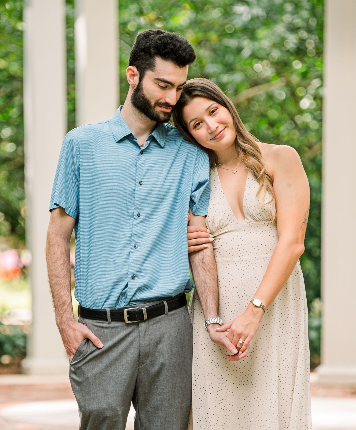 A couple poses outdoors near white columns, the woman in a beige maternity dress holding hands with a man in a blue shirt.