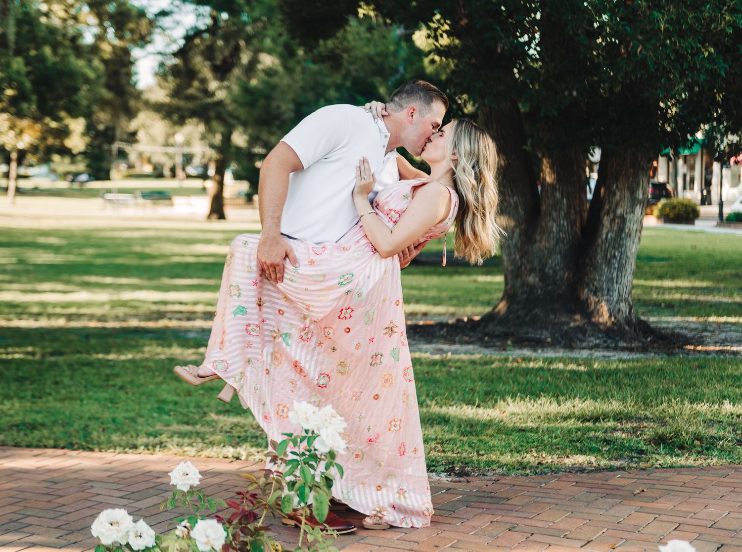 Couple sharing a romantic kiss in a lush green park, man dipping woman in a floral pink maxi dress.