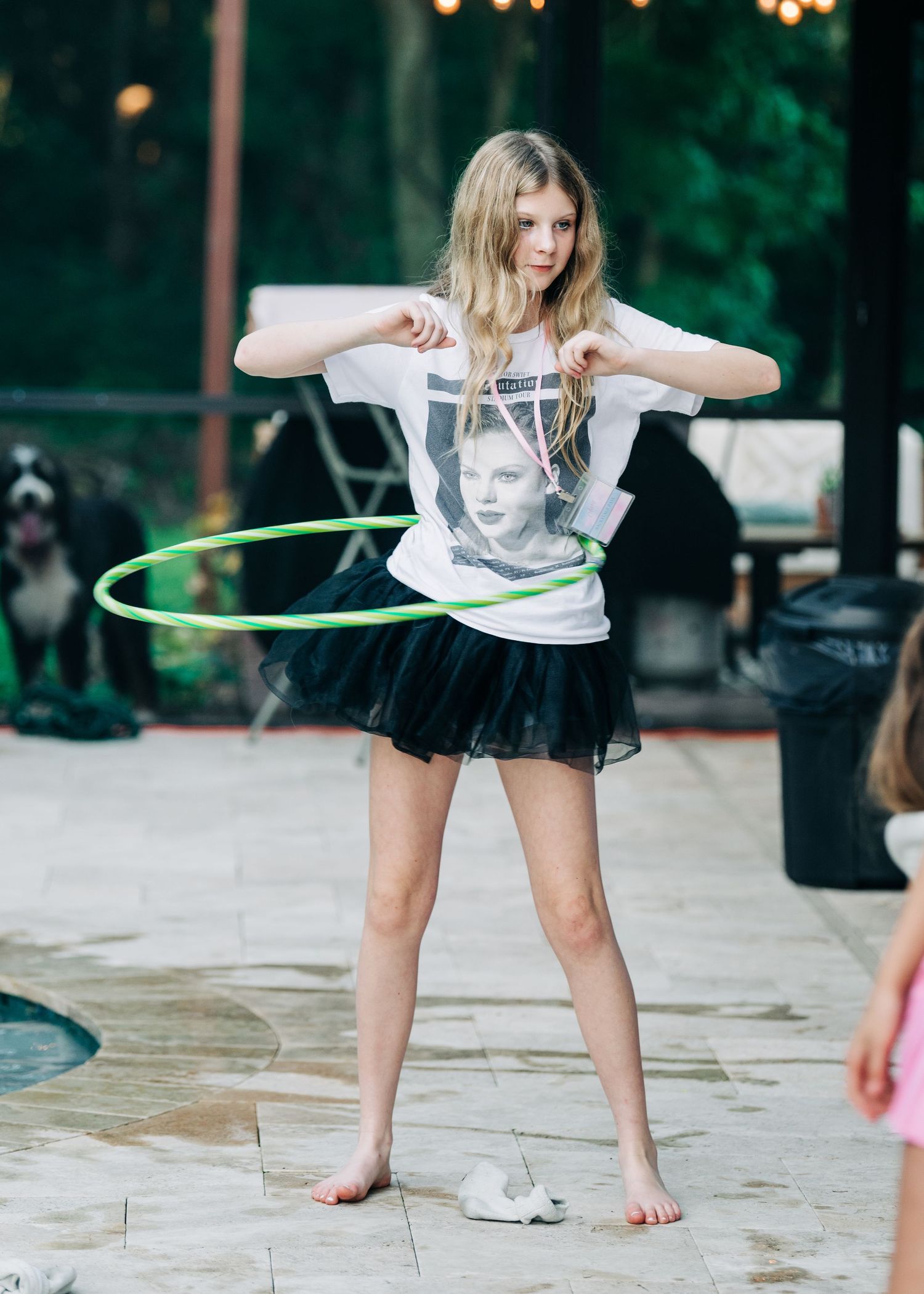 Young girl in white graphic tee and black tutu skirt hula hooping barefoot on a patio outdoors.