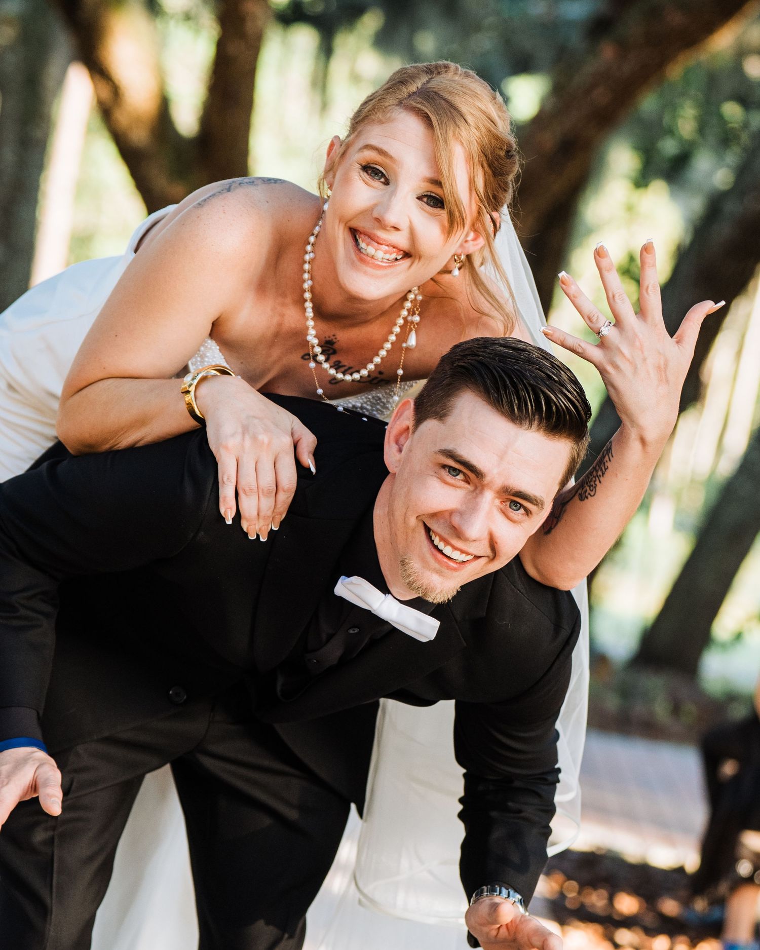 A smiling bride in white dress and pearl necklace piggybacks on a groom in black suit outdoors at a wedding.