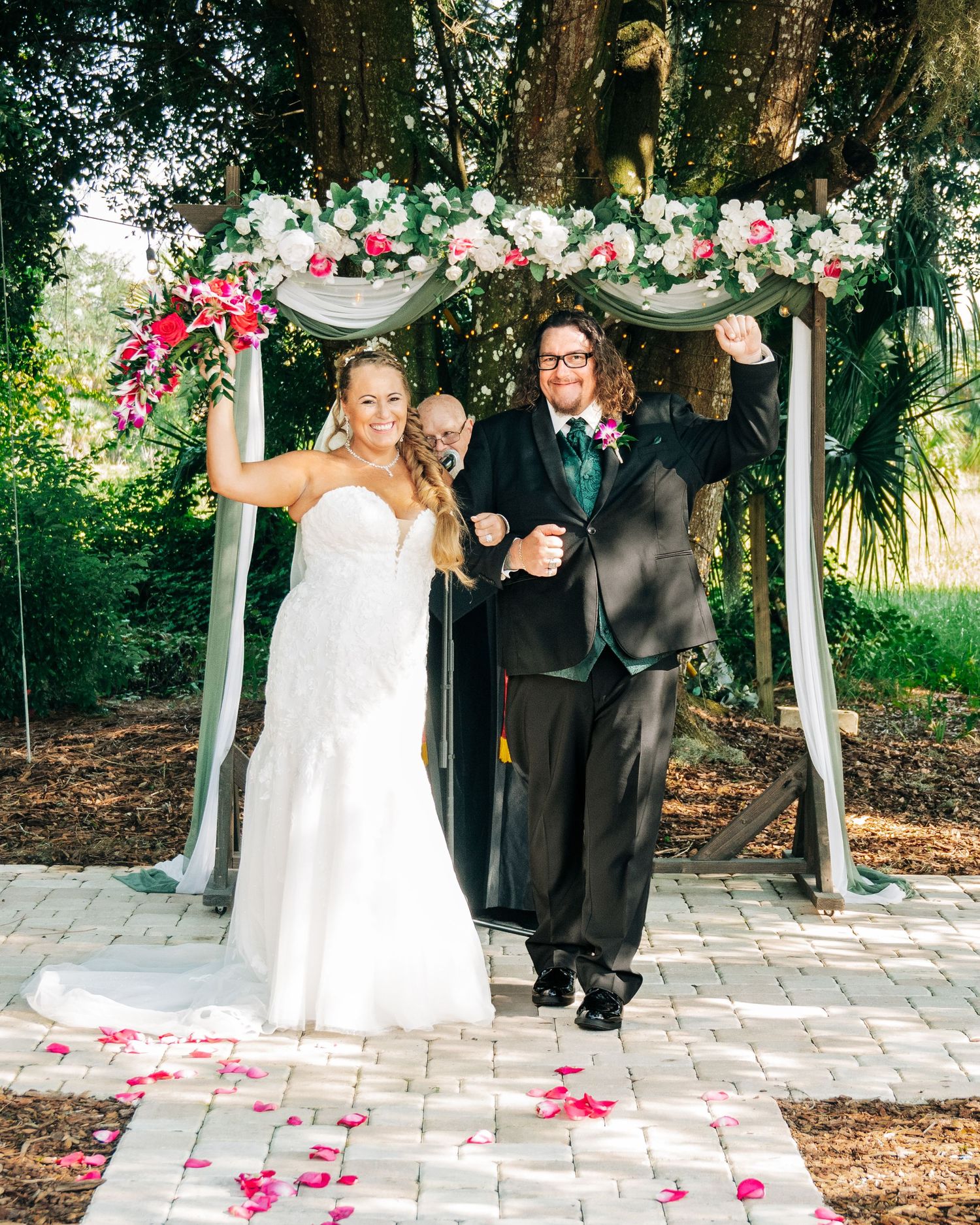 Joyful newlyweds celebrate under a floral arch with rose petals on a garden aisle after their outdoor wedding ceremony.