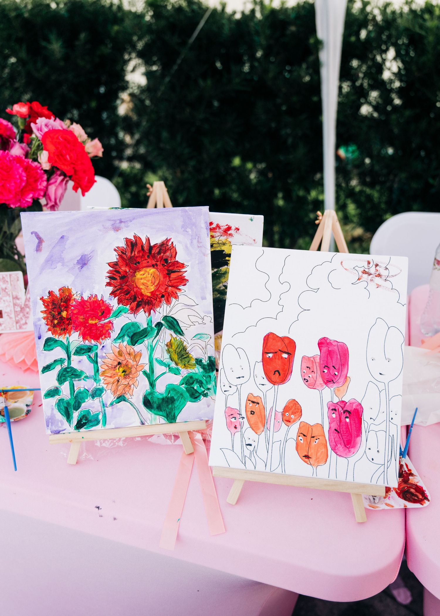 Two floral paintings on easels at an outdoor painting party, one with sunflowers and one with tulips featuring faces.