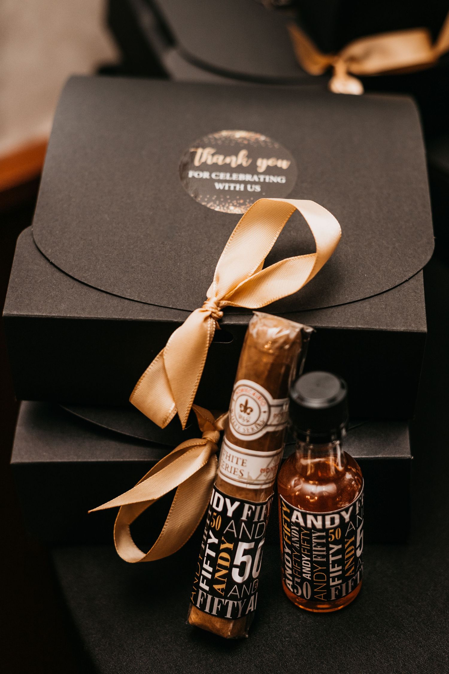 Black gift box with gold ribbon, cigar, and mini whiskey bottles labeled 50th birthday party favor.