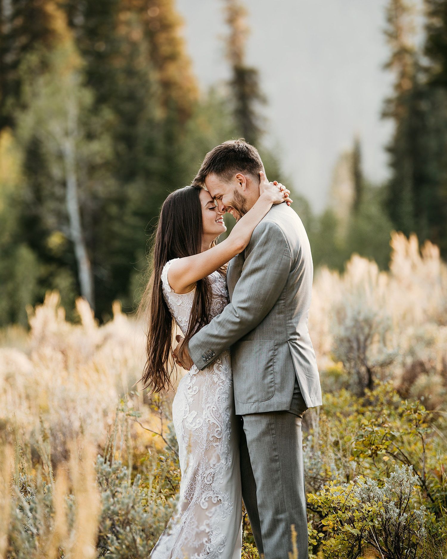 A couple in wedding attire embraces in a golden meadow surrounded by evergreen trees.