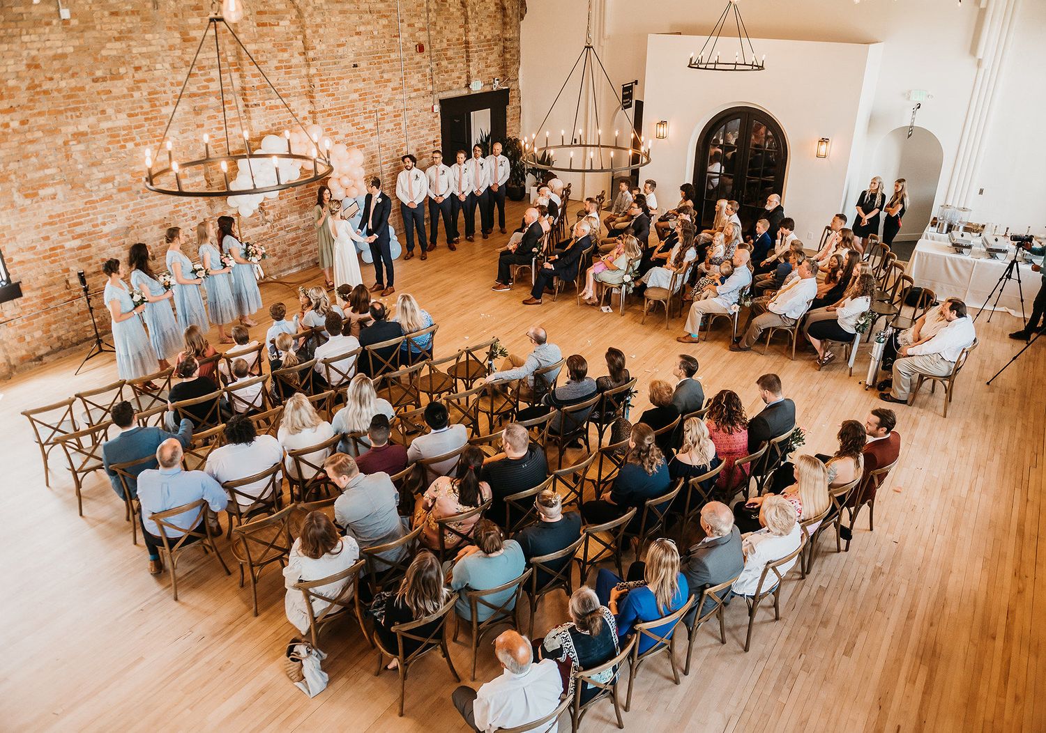 Aerial view of intimate indoor wedding ceremony with guests seated in rustic brick venue with wood floors.
