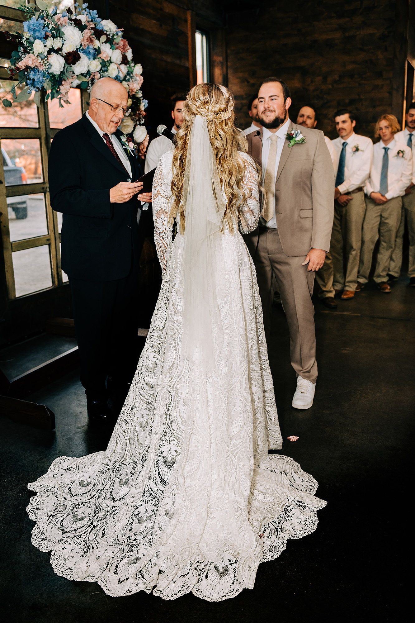 Bride in lace gown with long train stands at altar with groom in tan suit during indoor wedding ceremony.
