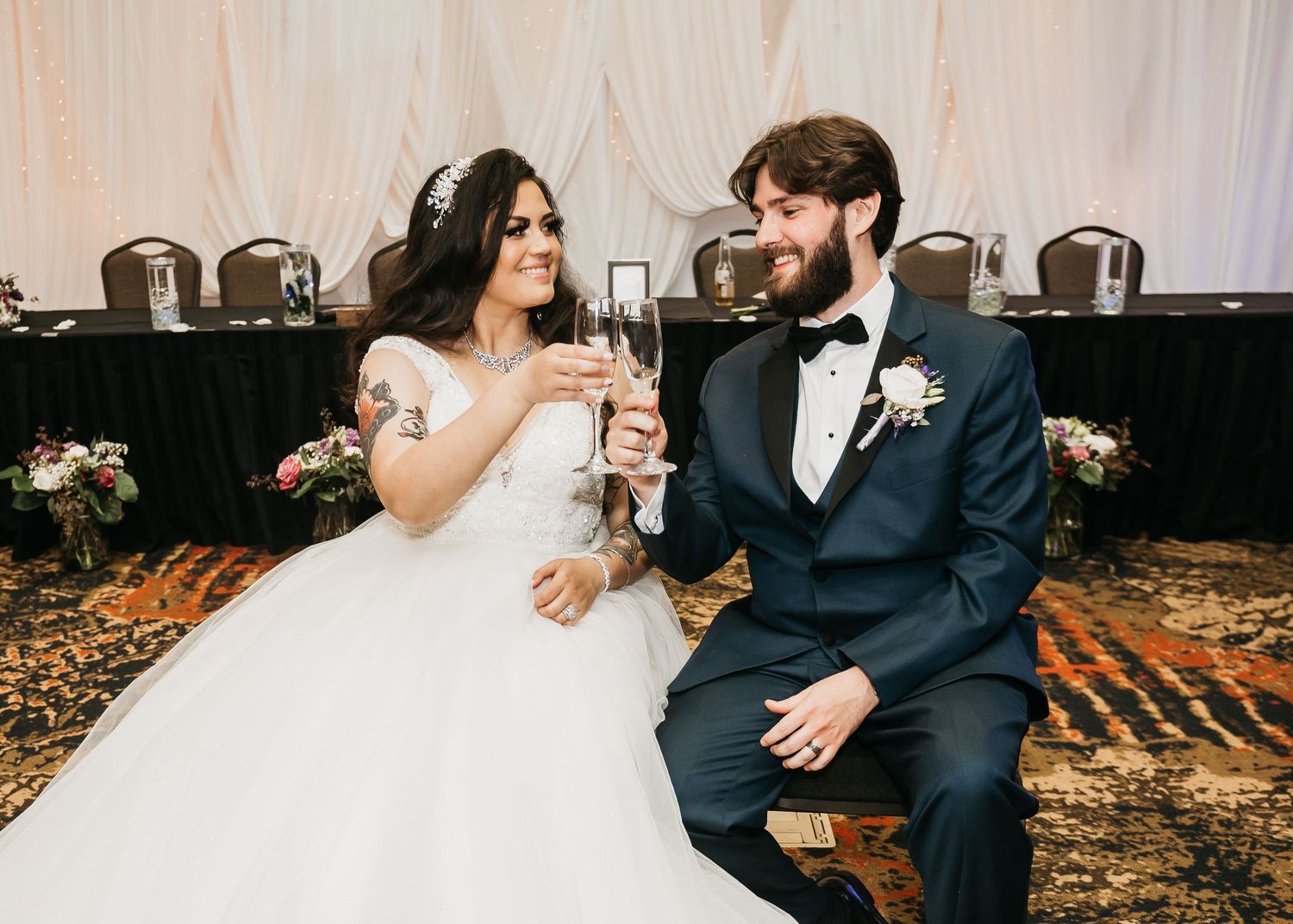 Bride and groom toasting with champagne glasses at wedding reception, bride in white gown, groom in navy tuxedo.