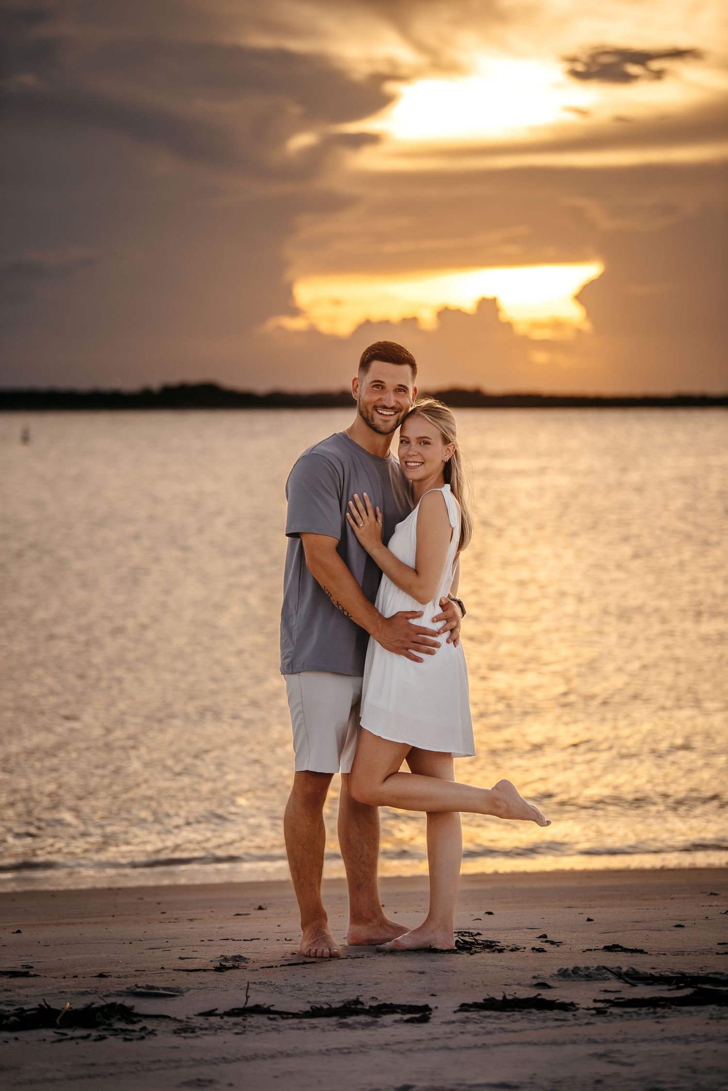 A couple embraces barefoot on a sandy beach at sunset, smiling warmly with golden skies and calm water behind them.
