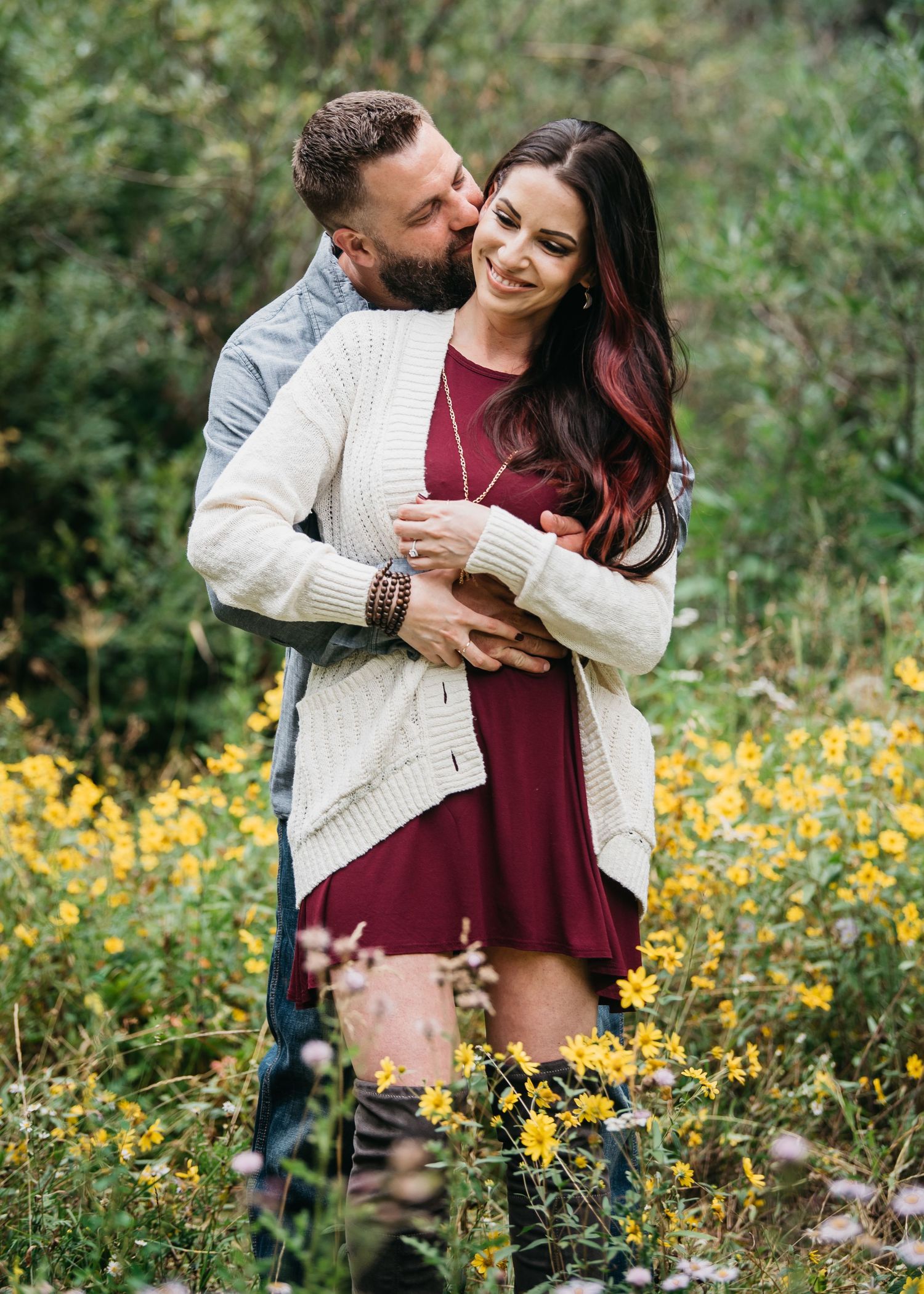 Couple embracing in a wildflower field, woman in burgundy dress and cream cardigan, man kissing her cheek.