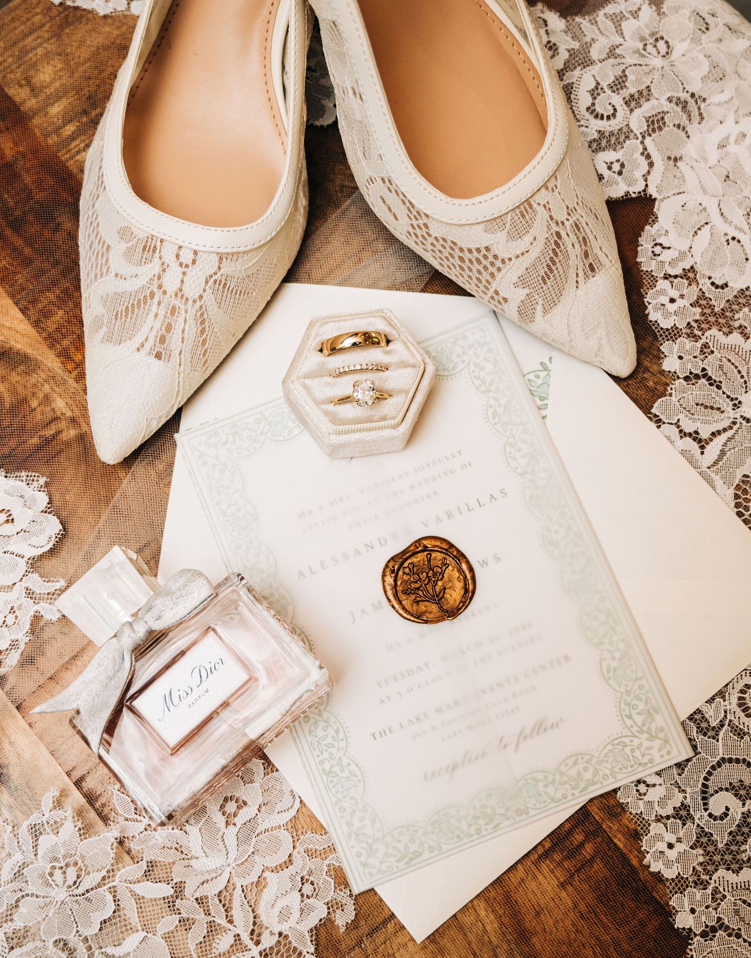 Elegant wedding flat lay with lace heels, gold rings, Miss Dior perfume, wax seal invitation on rustic wood.