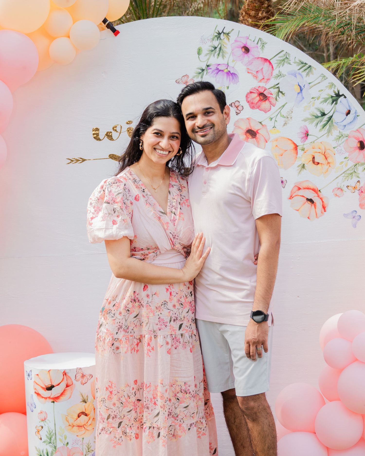 Smiling couple posing together at a floral baby shower with pink balloons and a flower backdrop.