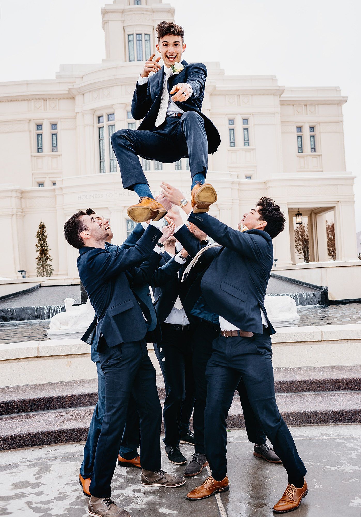 Groomsmen in navy suits tossing groom into the air outside elegant white wedding venue.
