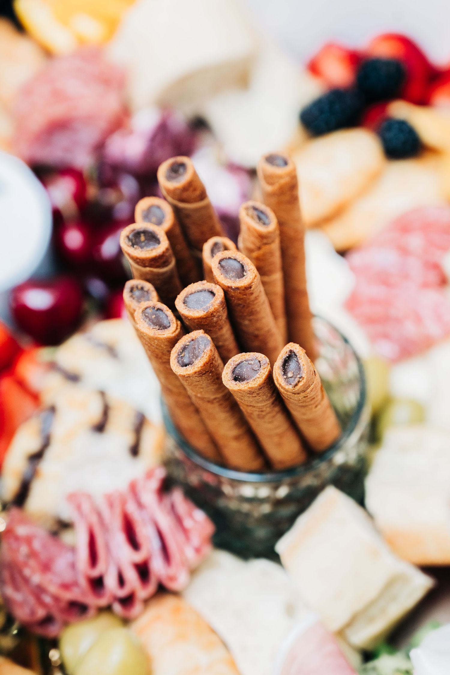 Chocolate wafer rolls in a glass on a charcuterie board with cheese, salami, cherries, and berries.