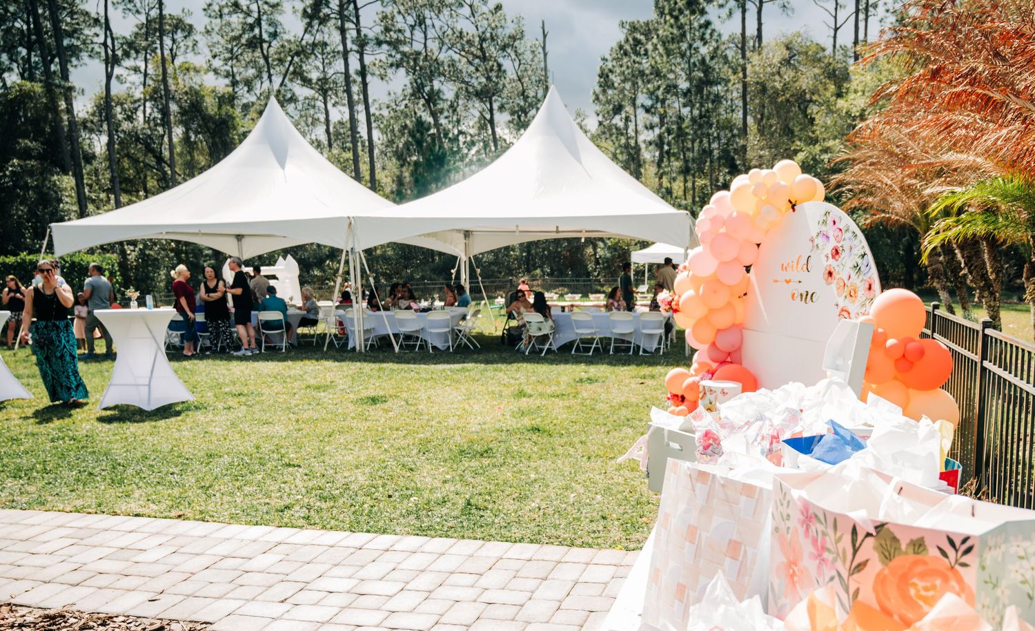 Outdoor party setup with white canopy tents, balloon decorations, and guests seated at tables on a sunny day.