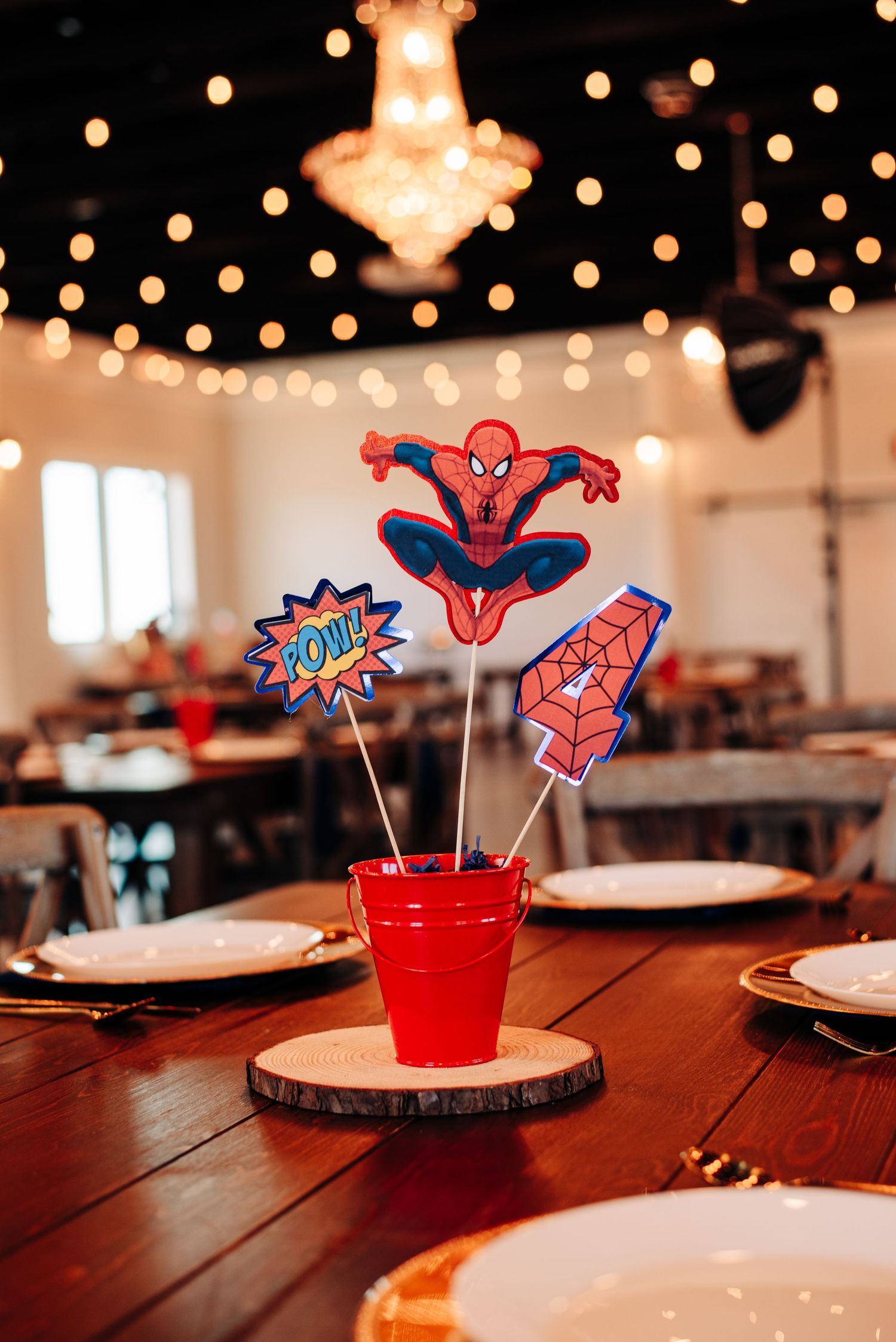 Spider-Man themed party centerpiece in red bucket with POW sign on wooden table, string lights and chandelier in background.