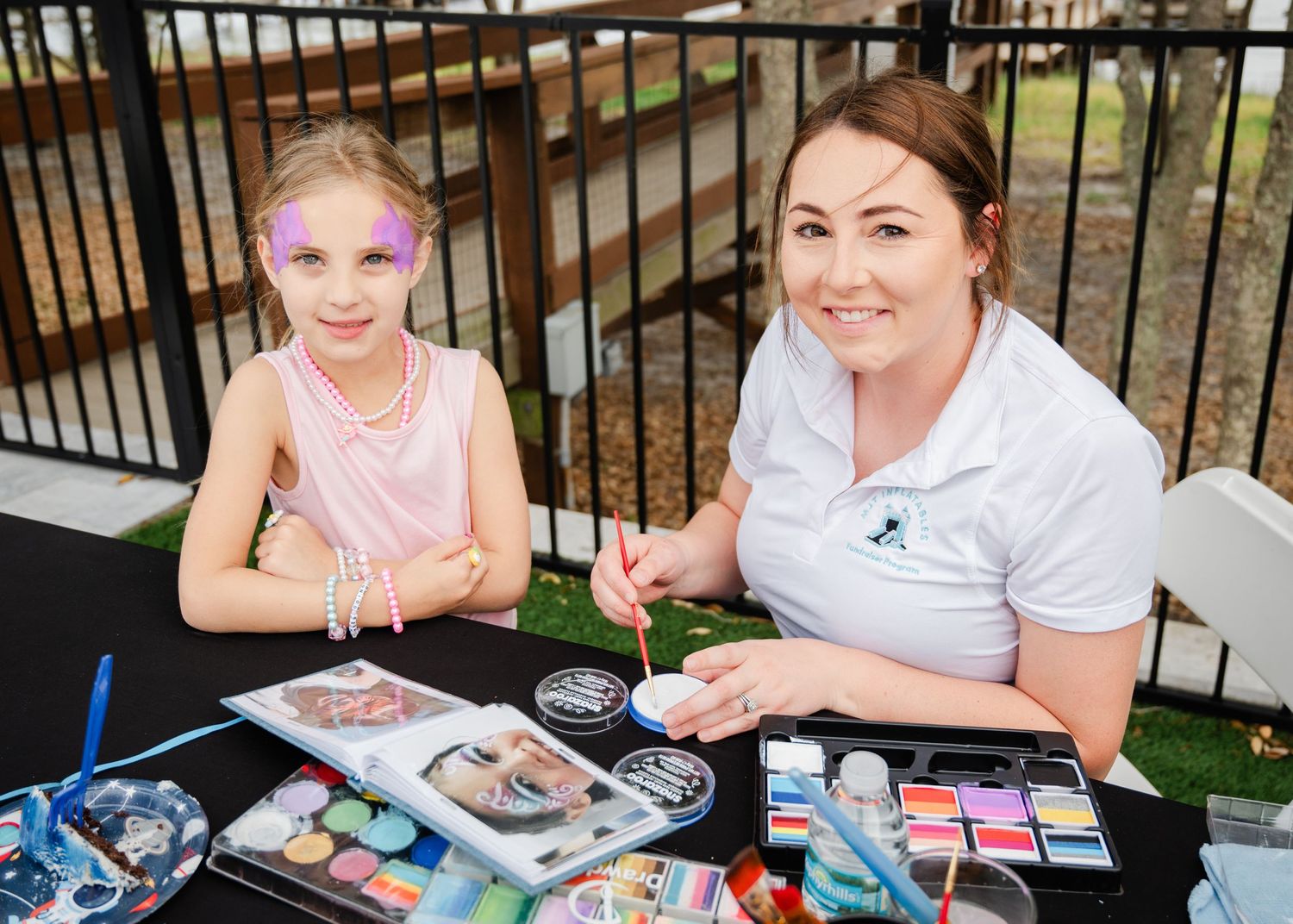 A face painter applies colorful designs on a young girl's face at an outdoor event table with supplies.