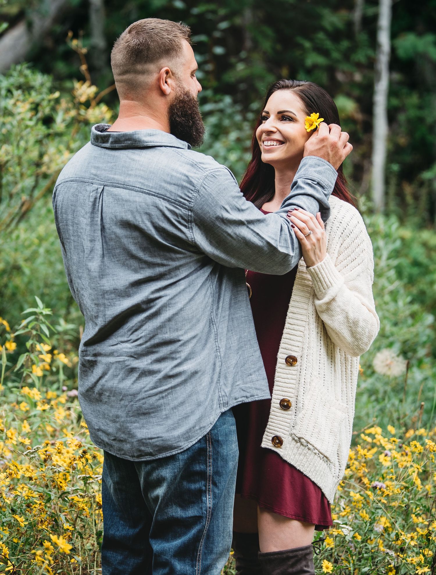 A bearded man places a yellow flower in a smiling woman's hair outdoors among wildflowers.