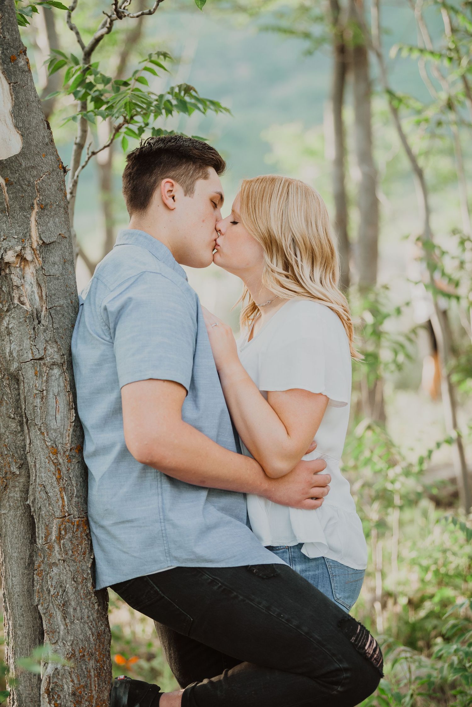 A couple shares a romantic kiss leaning against a tree in a lush green forest during an outdoor engagement photo session.