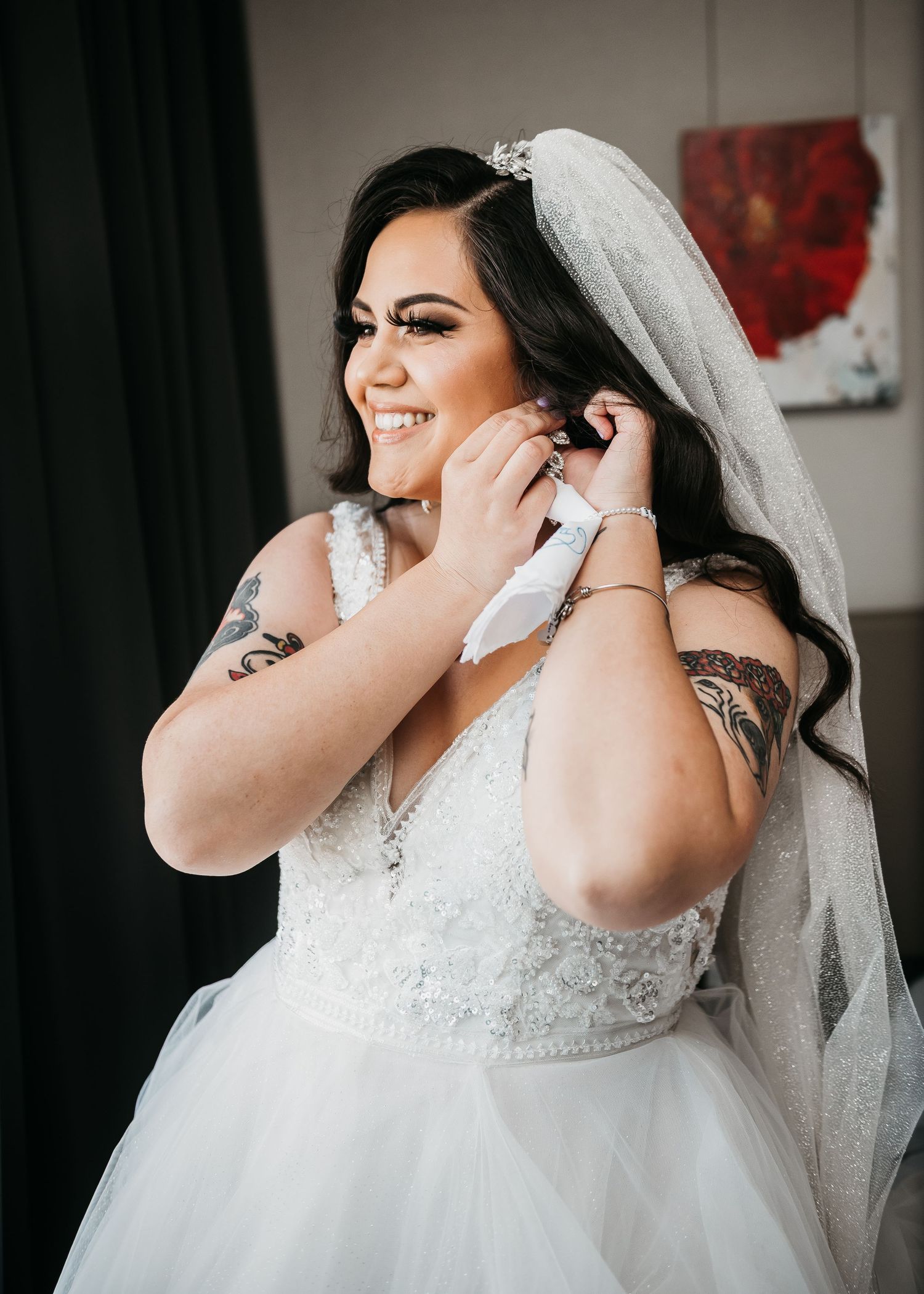 Smiling bride in white lace ballgown and veil adjusting earring, showcasing tattoos on her arms.