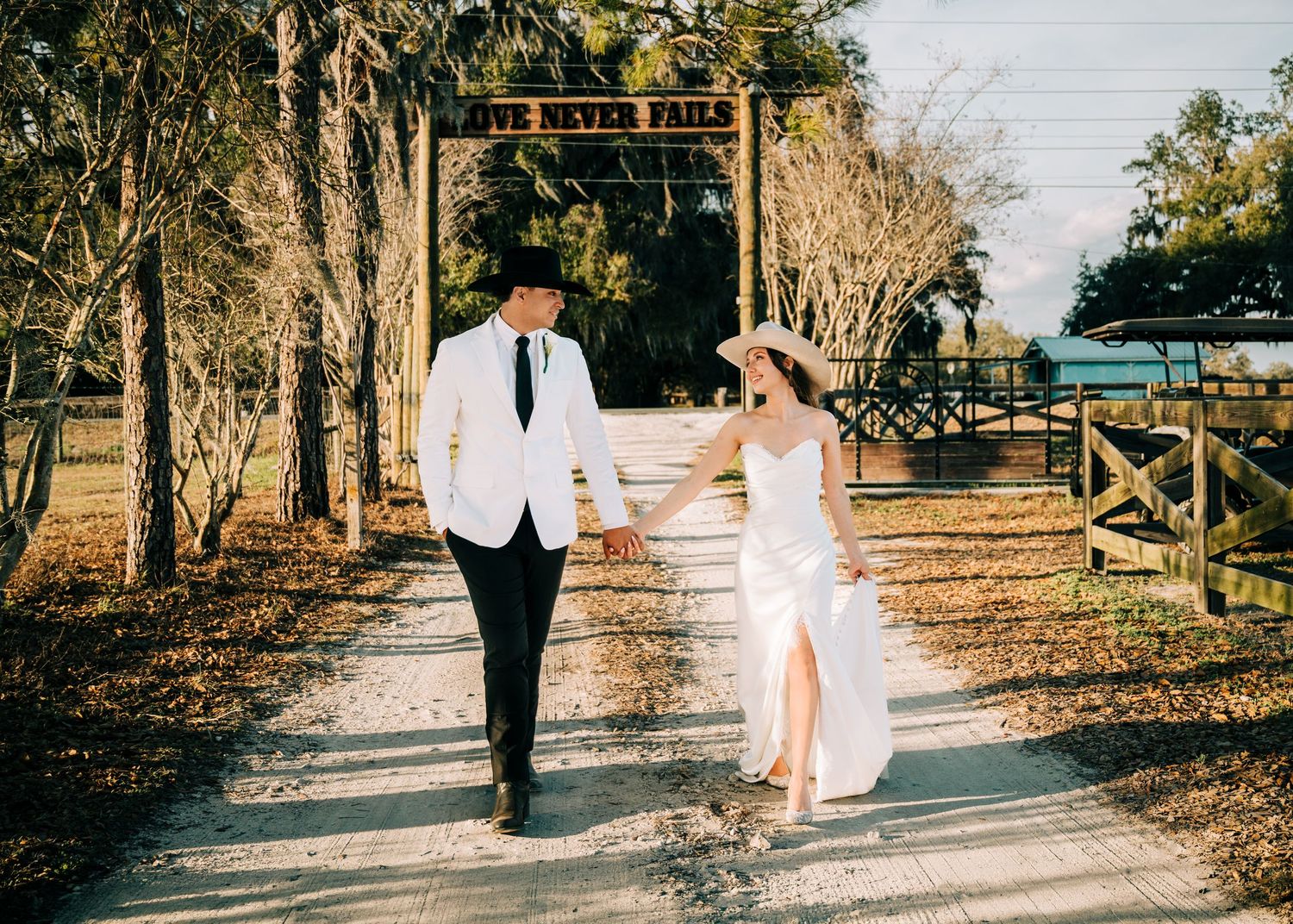 A bride and groom walk hand-in-hand down a rustic path beneath a Love Never Fails sign at an outdoor wedding venue.