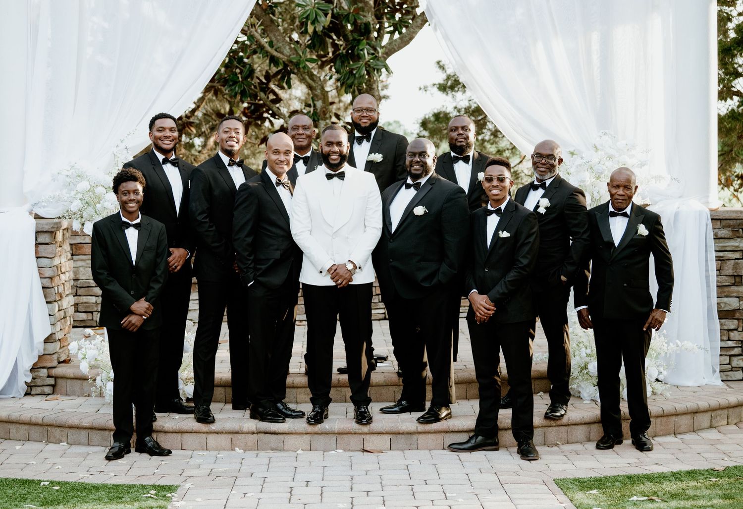 Groom in white tuxedo jacket poses with groomsmen in black tuxedos at outdoor wedding ceremony.