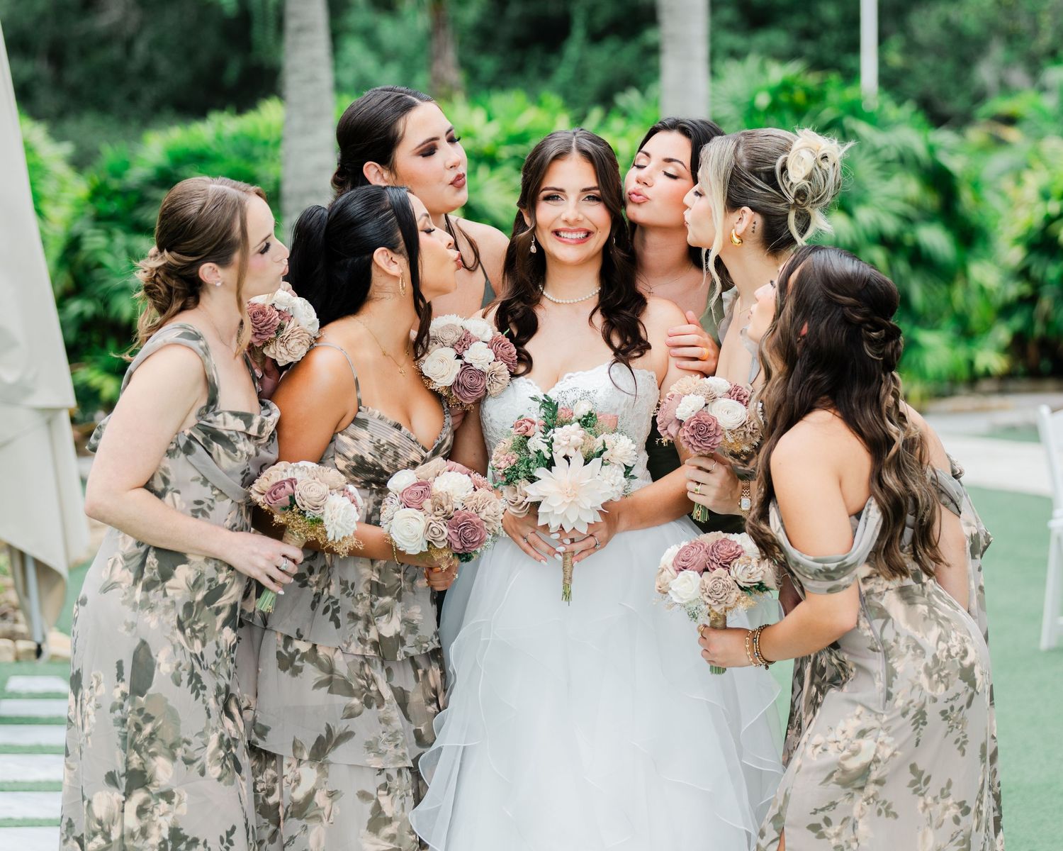 Bride surrounded by bridesmaids in floral dresses holding bouquets, sharing a joyful group moment outdoors.
