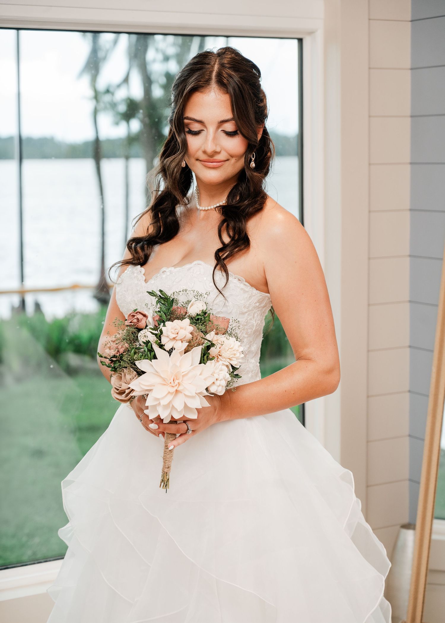 Bride in strapless lace ballgown holds blush dahlia bouquet, standing by window with lake view.