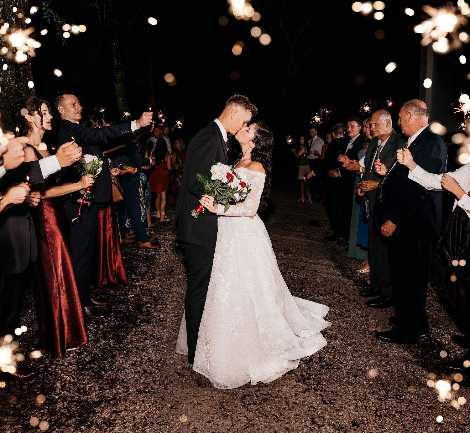 Bride and groom kissing during sparkler exit at night wedding ceremony with guests lining the path.