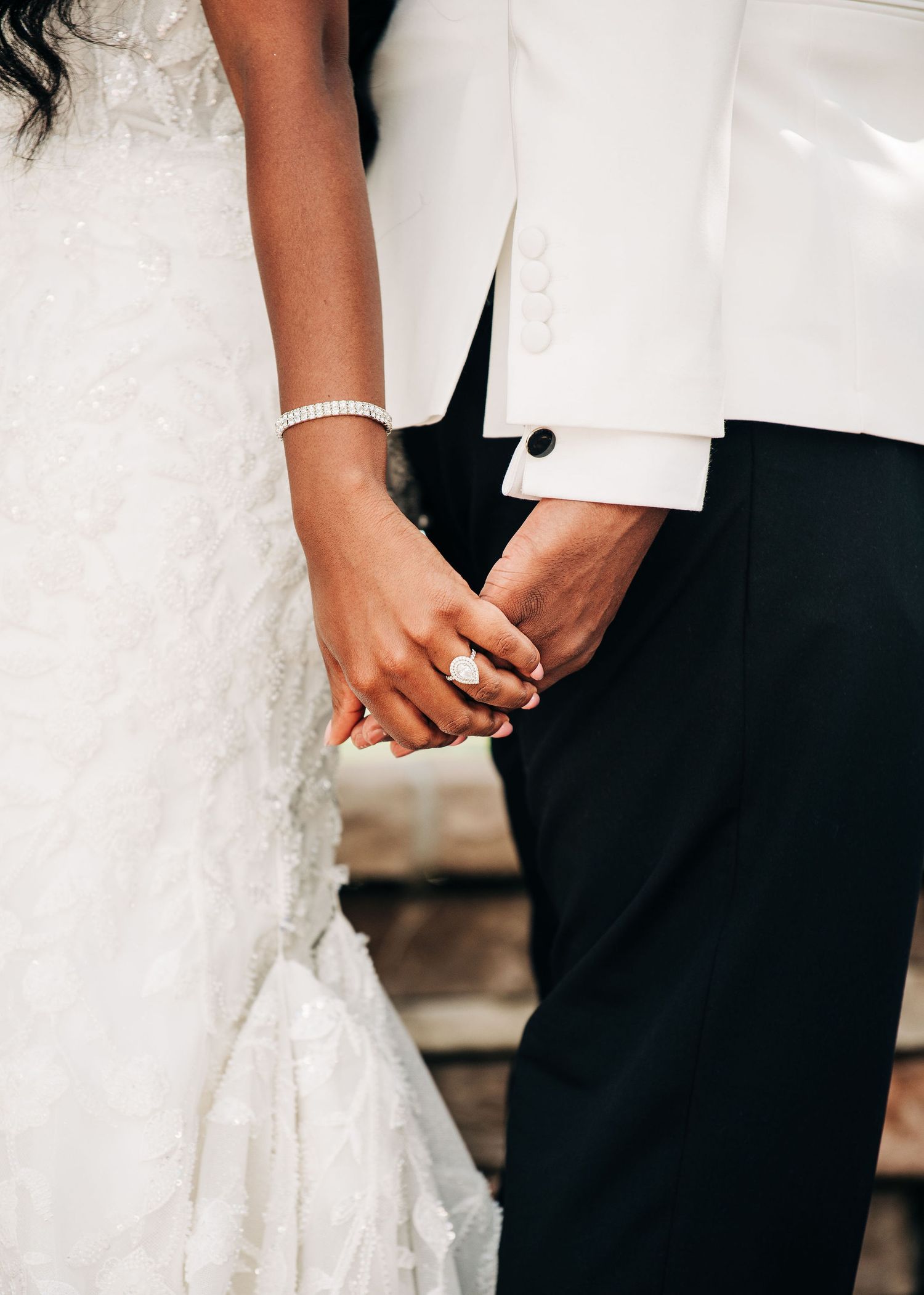 Bride and groom holding hands, showcasing diamond engagement ring and bracelet, wedding attire close-up.