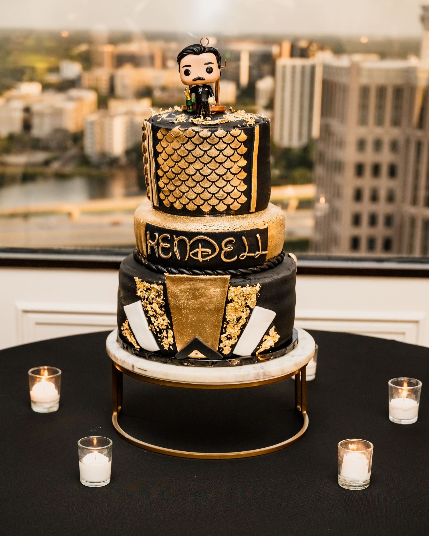 Black and gold three-tier wedding cake with Funko Pop topper, reading Rendell, displayed on rooftop venue with city views.