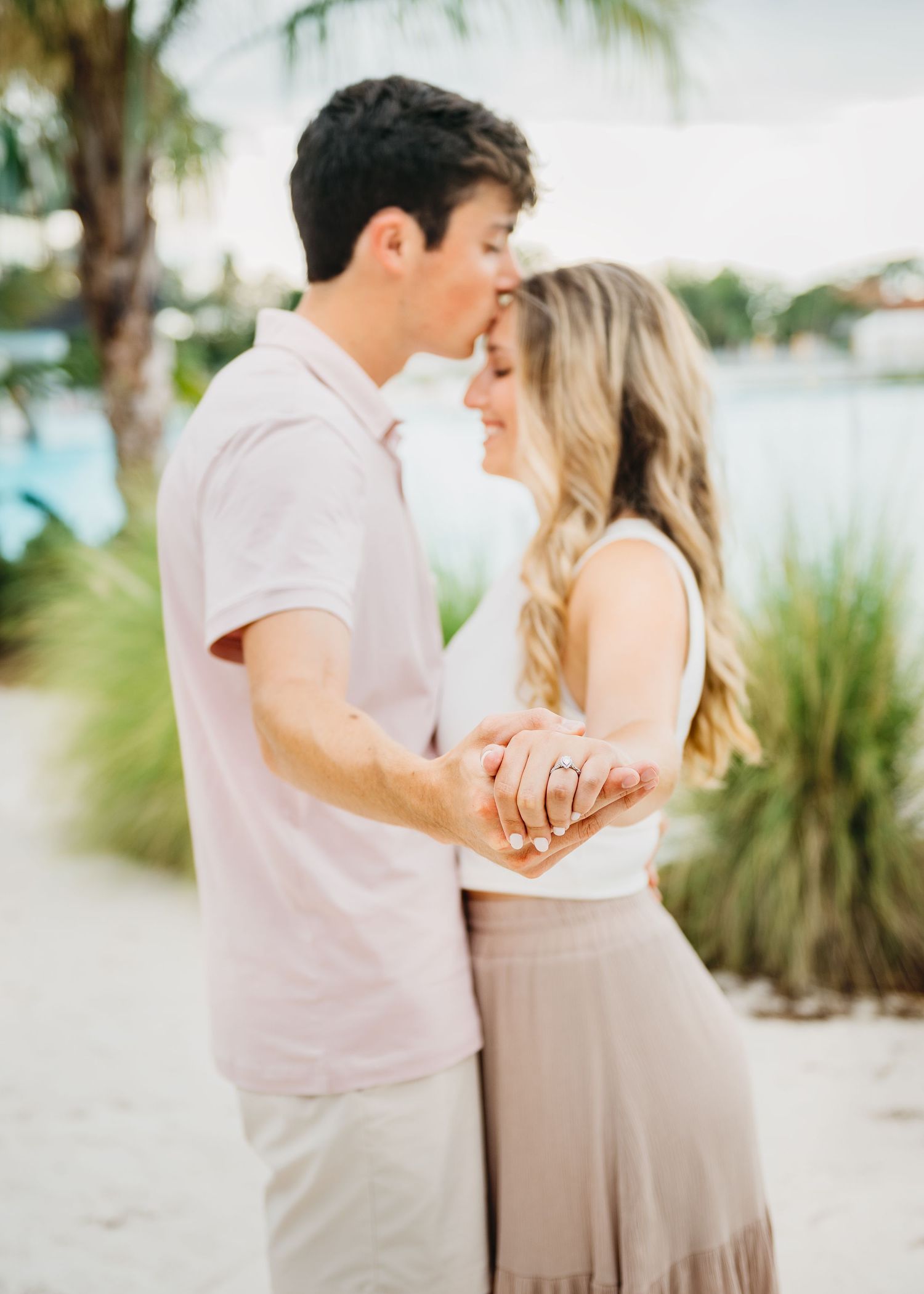 Couple sharing a romantic kiss outdoors near a beach, holding hands and showing an engagement ring.