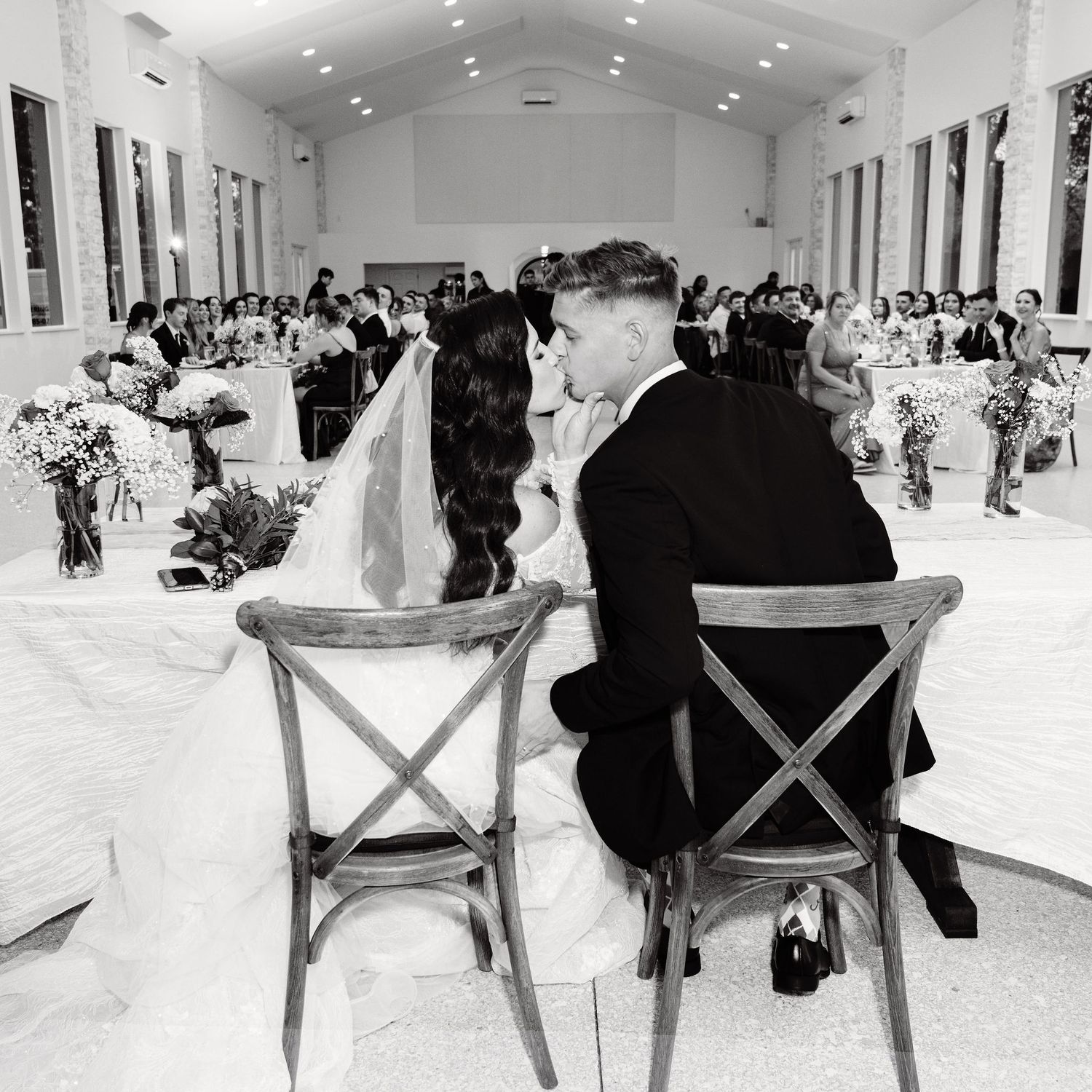 Black and white photo of bride and groom seated at sweetheart table during elegant wedding reception.