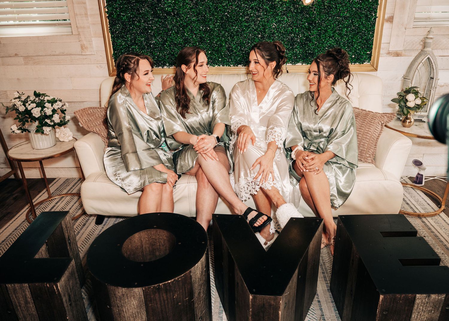 Bride and bridesmaids in satin robes sitting together on a couch, laughing, with a LOVE sign in foreground.