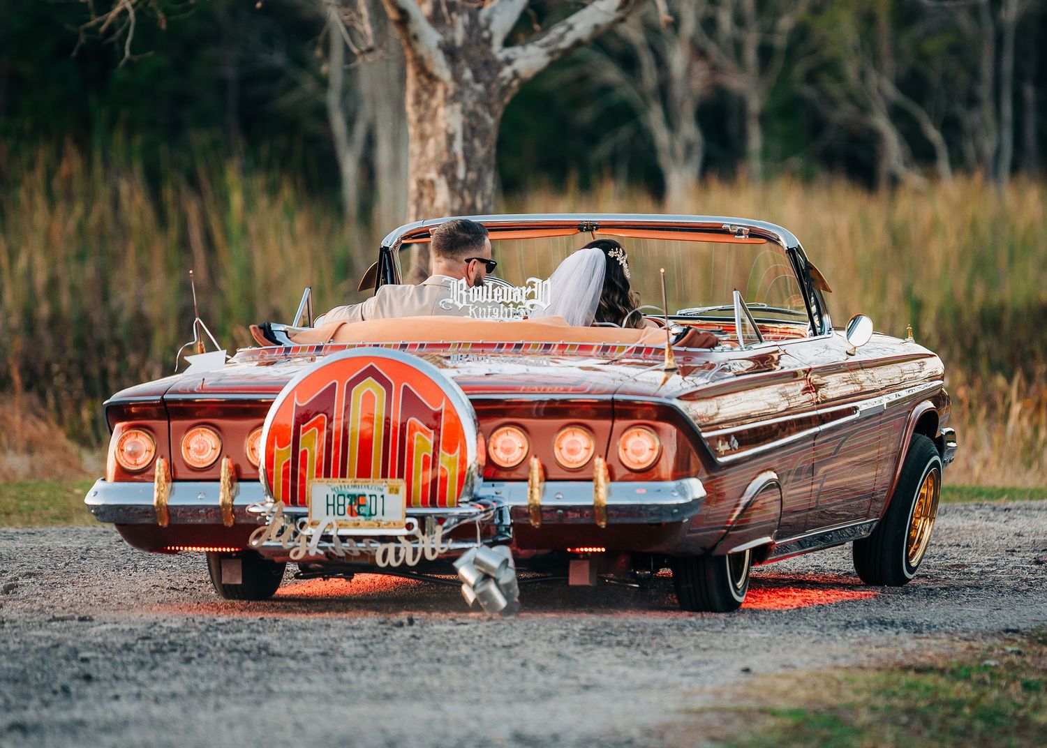 Vintage wooden convertible lowrider car with glowing taillights at a wedding, bride and groom seated inside, parked on gravel.