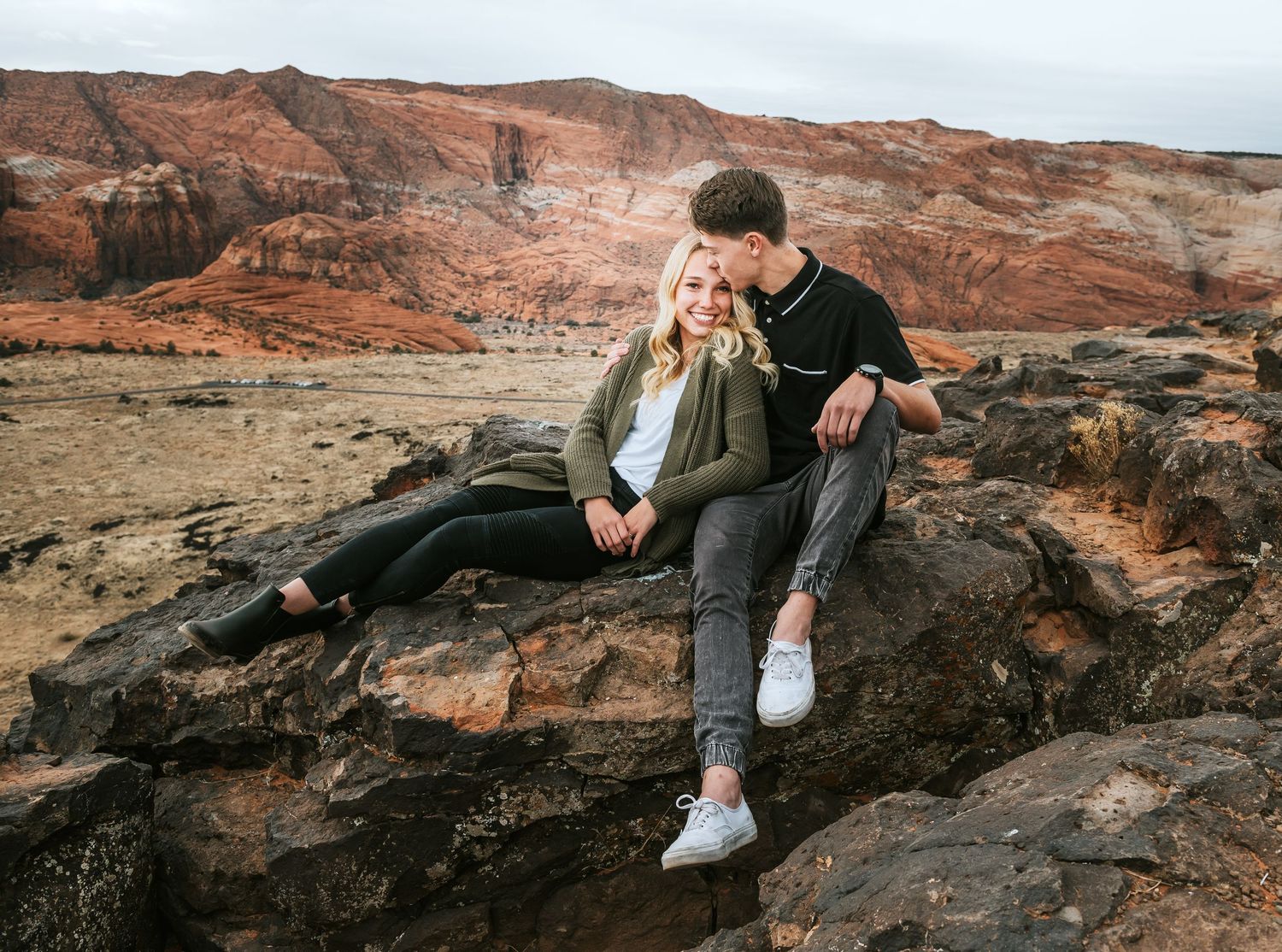 Couple sitting on rocks in a desert landscape with red rock formations in the background.