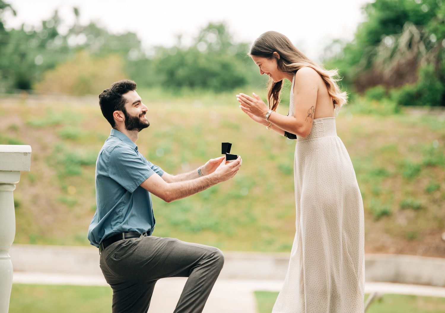 Man kneeling on one knee proposing to woman outdoors, holding open ring box during romantic marriage proposal.