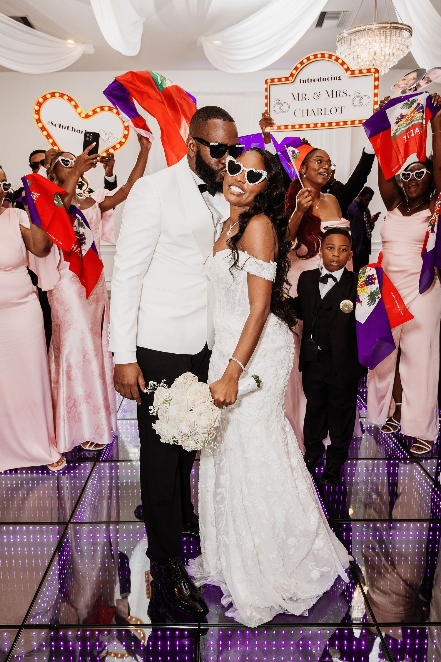 Newlyweds Mr. and Mrs. Charlot pose on a mirrored dance floor surrounded by guests waving flags at their wedding reception.