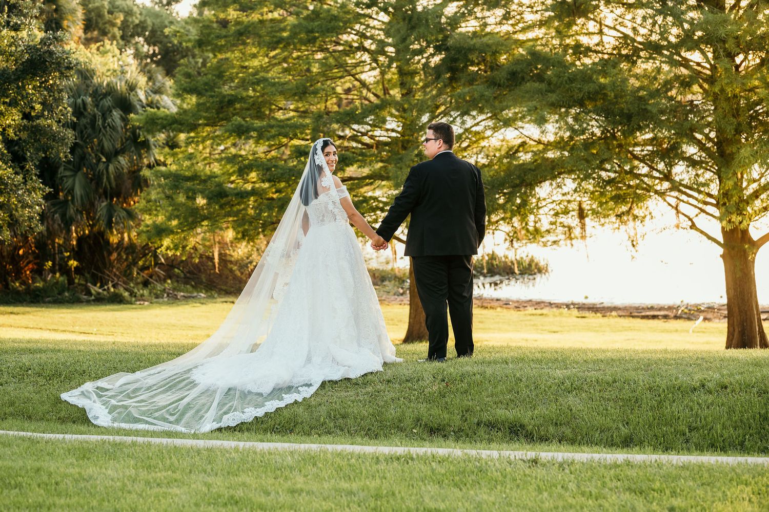 Bride in white gown with long veil holds hands with groom in black suit walking on green lawn near trees.