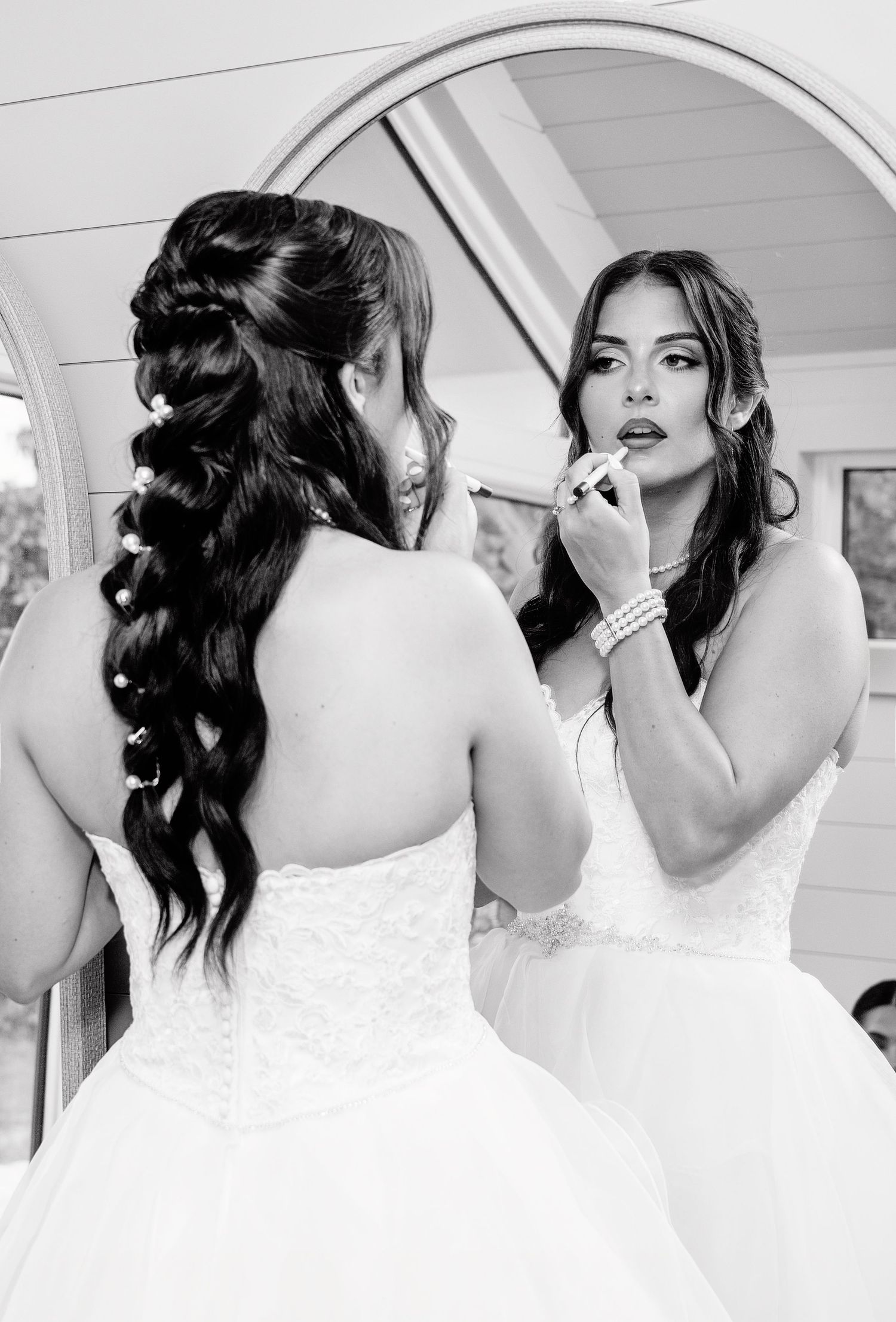 Bride applying lipstick while reflected in a round mirror, wearing a white gown with an elegant braided hairstyle.
