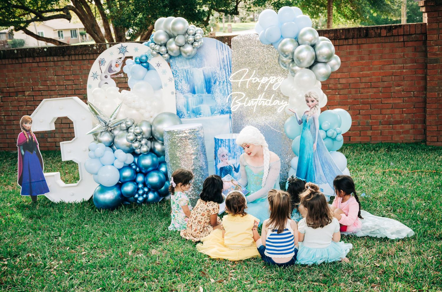 Frozen-themed birthday party with balloon arch, number 3, Elsa character, and children gathered on grass.