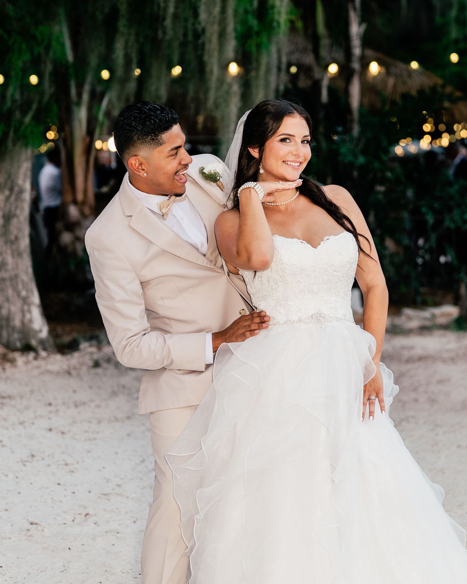 A couple poses outdoors at their wedding, groom in beige suit, bride in strapless white ballgown with veil, string lights behind.