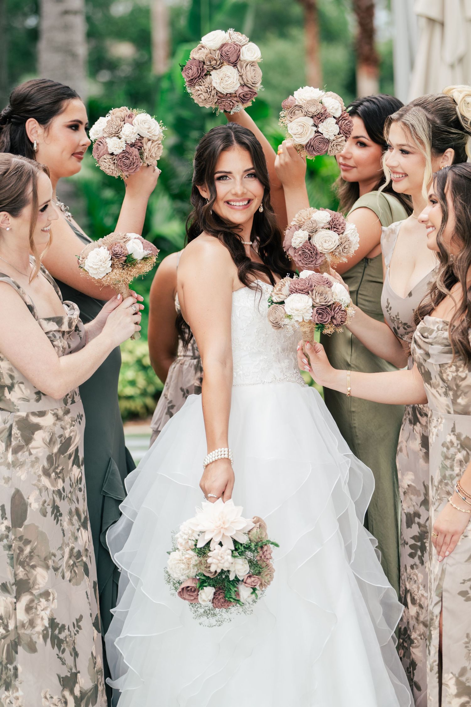 Bride in white gown poses with bridesmaids holding floral bouquets aloft at an outdoor wedding celebration.