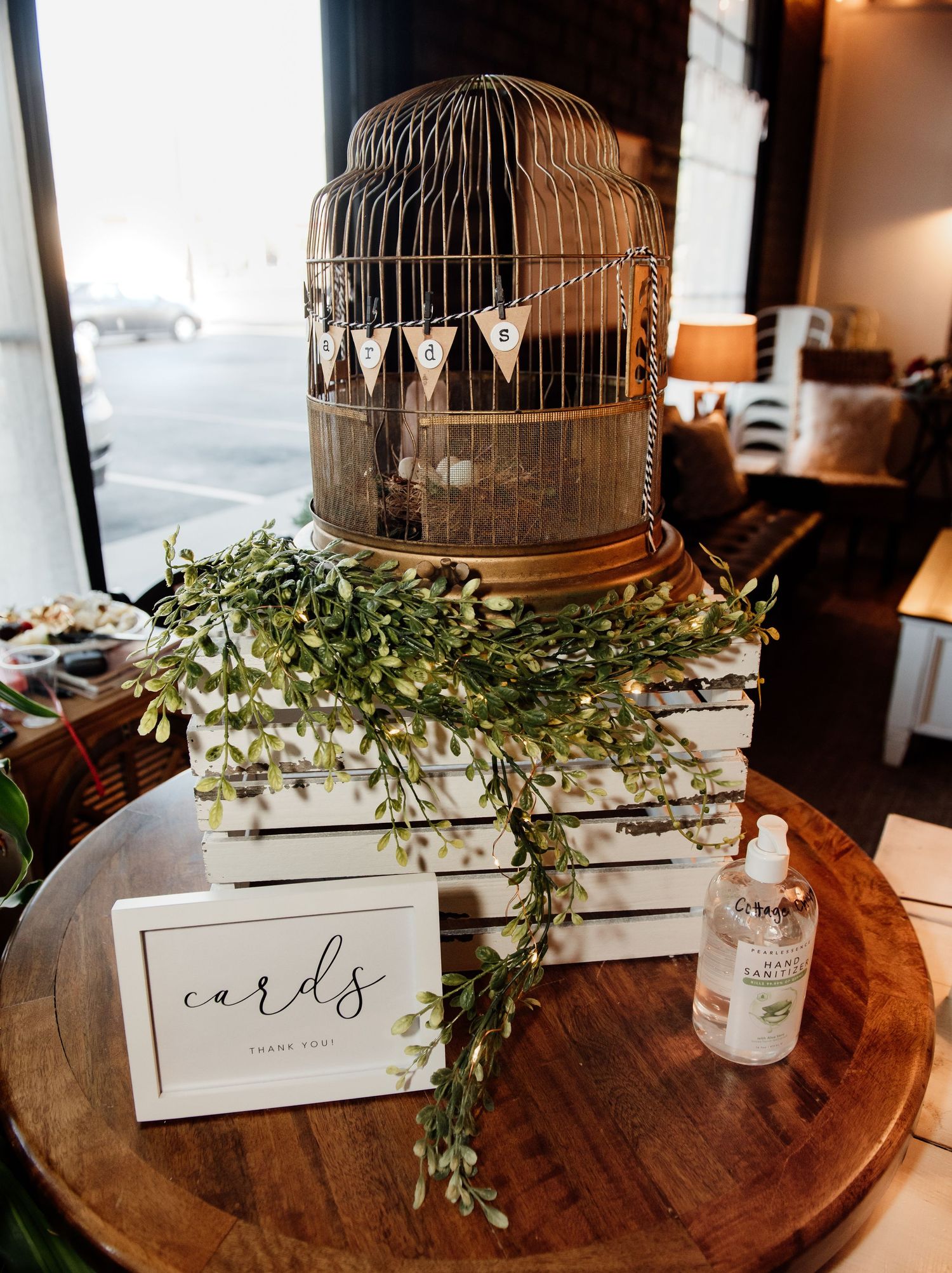 Rustic wedding card box display with gold birdcage, white crate, greenery garland, and hand sanitizer on wooden table.