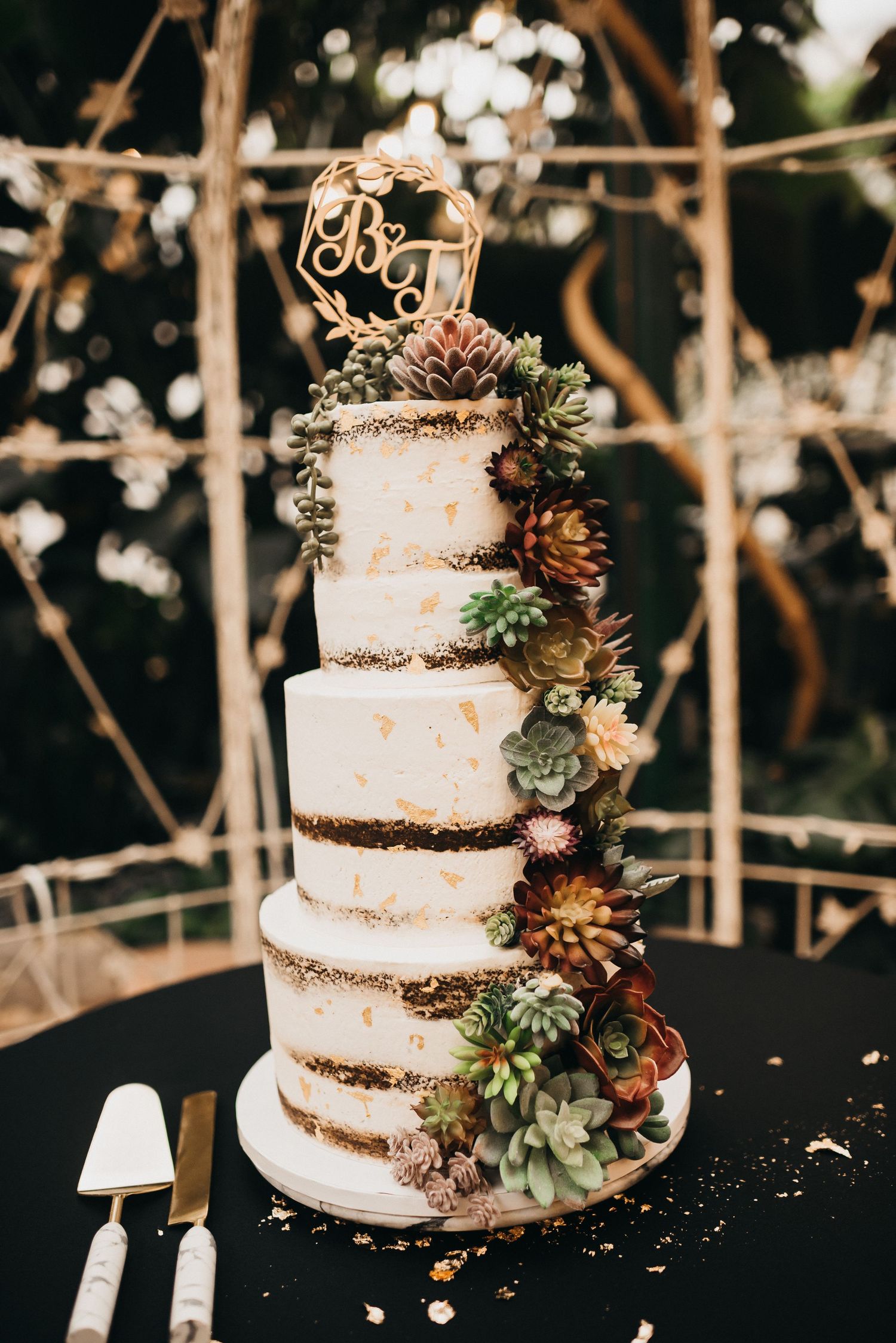 Four-tier white wedding cake with gold leaf accents, cascading succulents, and monogram topper on black tablecloth.