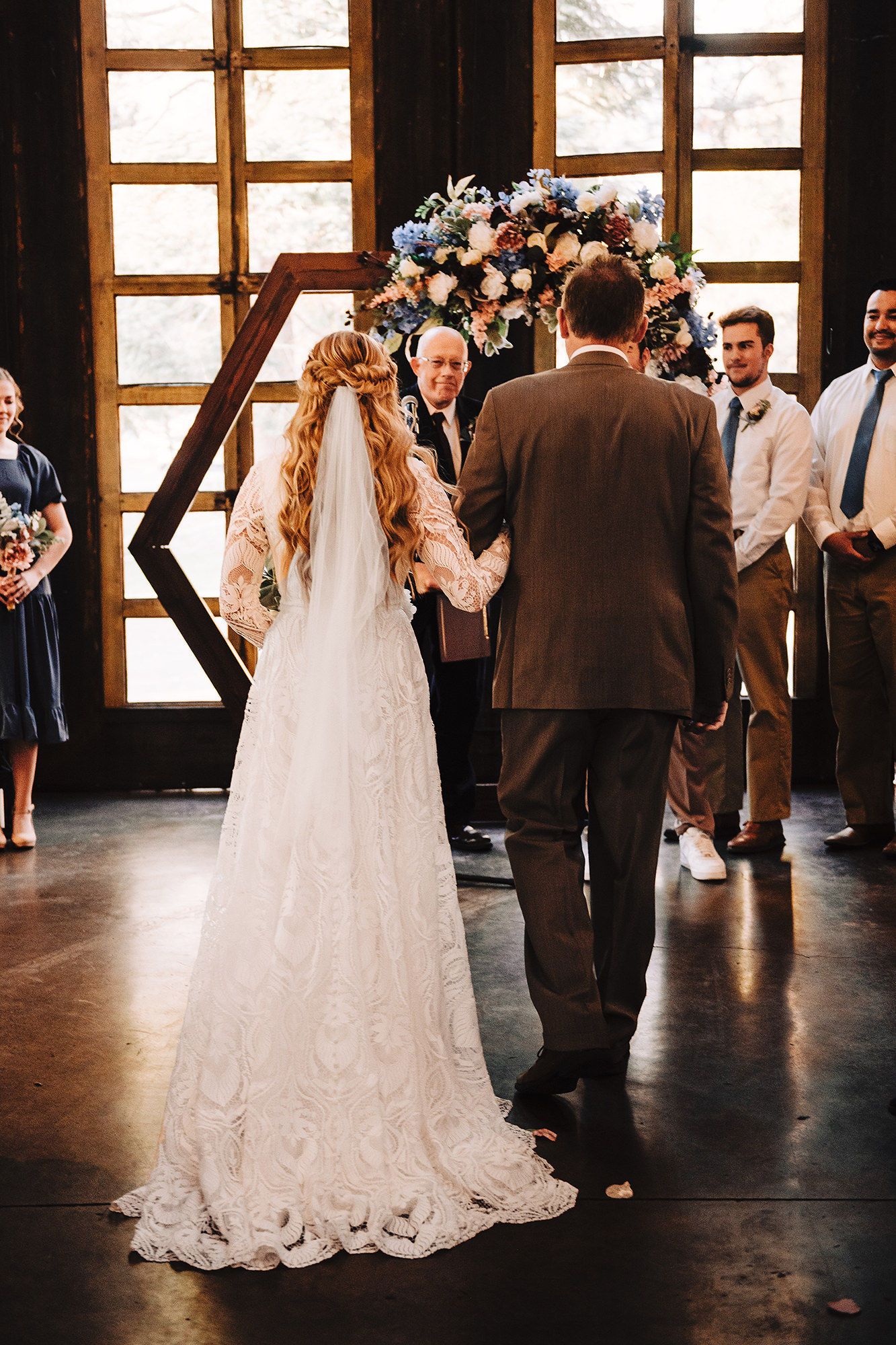 Bride and groom hold hands at indoor wedding altar adorned with blue and pink floral arch and geometric wooden backdrop.