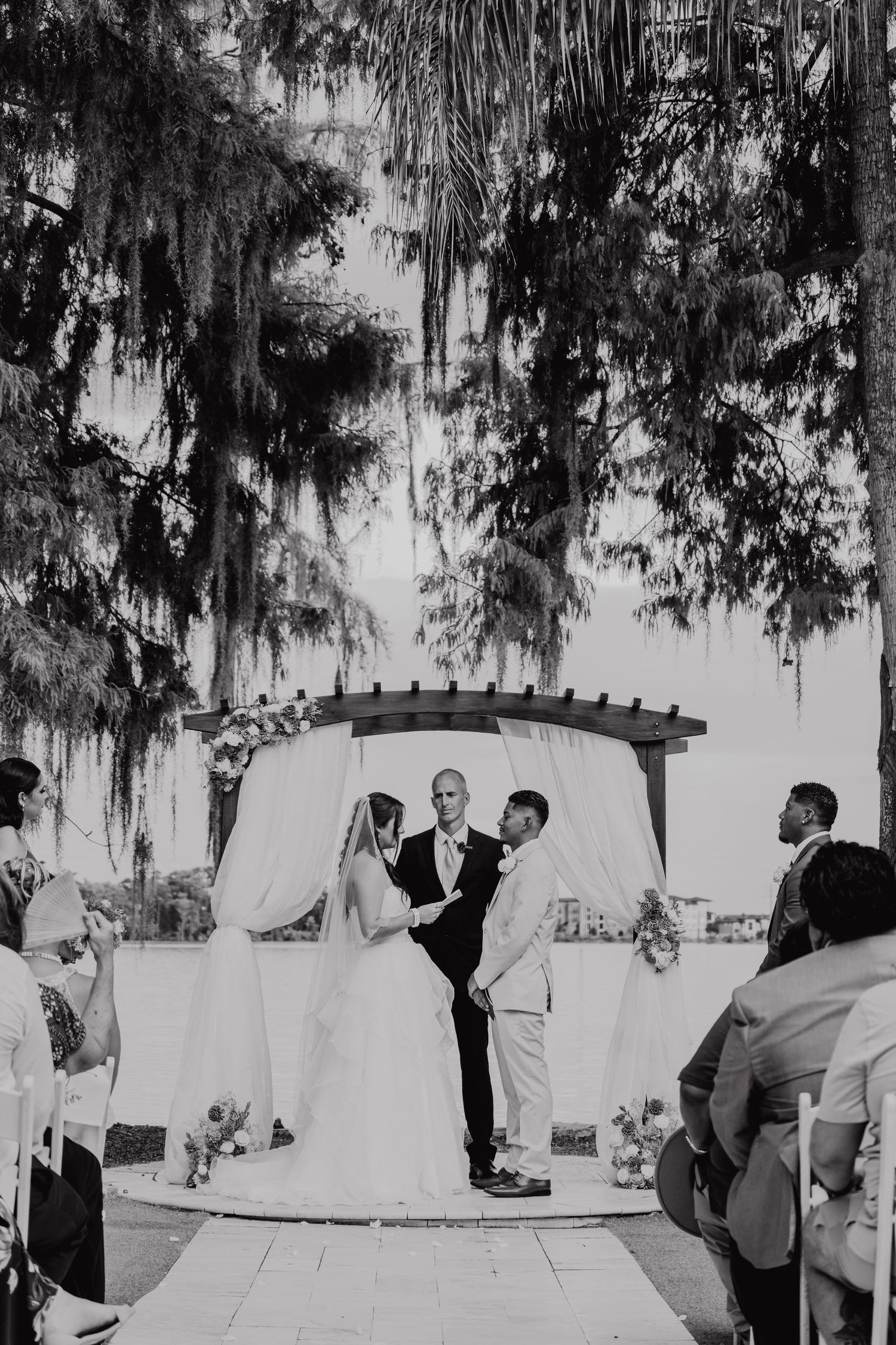 Black and white outdoor wedding ceremony with couple exchanging vows under a pergola draped in white fabric and lush trees.