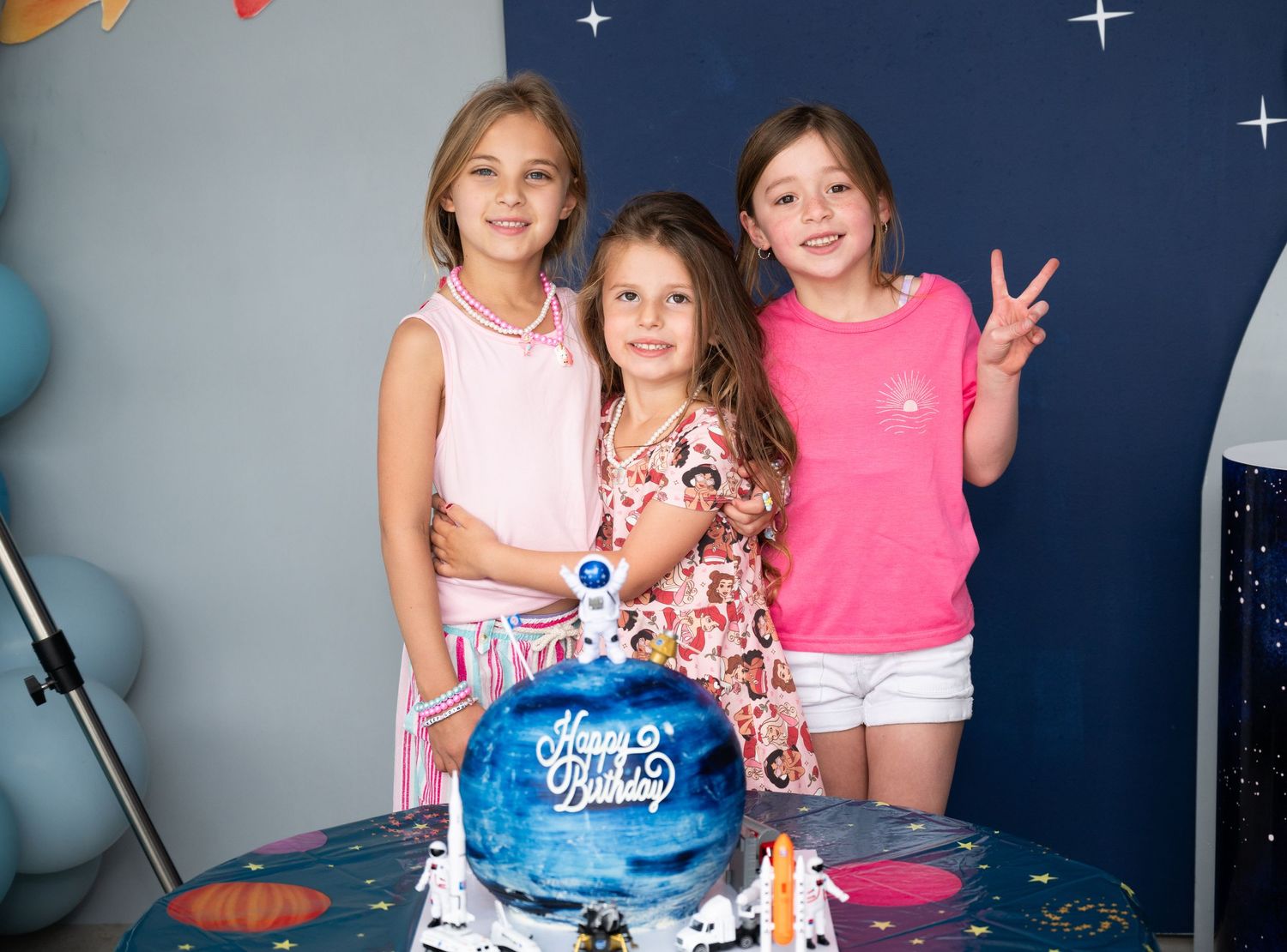 Three young girls smile together at a space-themed birthday party with a galaxy cake on the table.