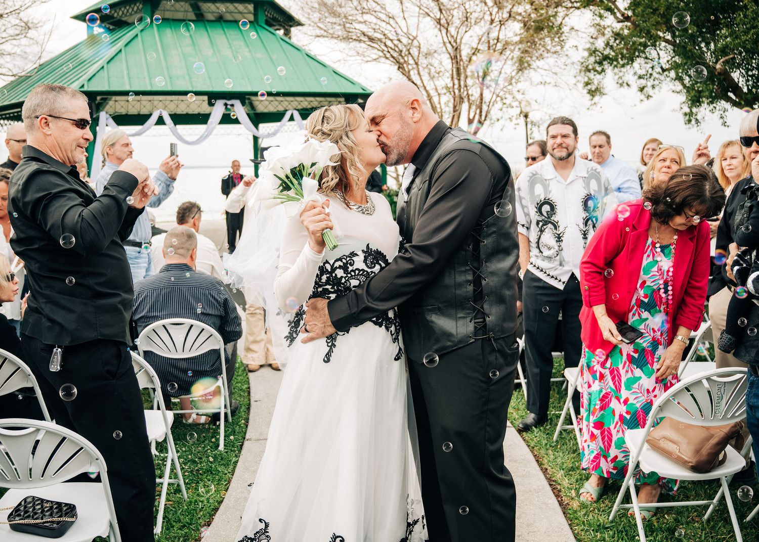 Bride and groom share first kiss at outdoor wedding ceremony near green gazebo with guests seated in white chairs.