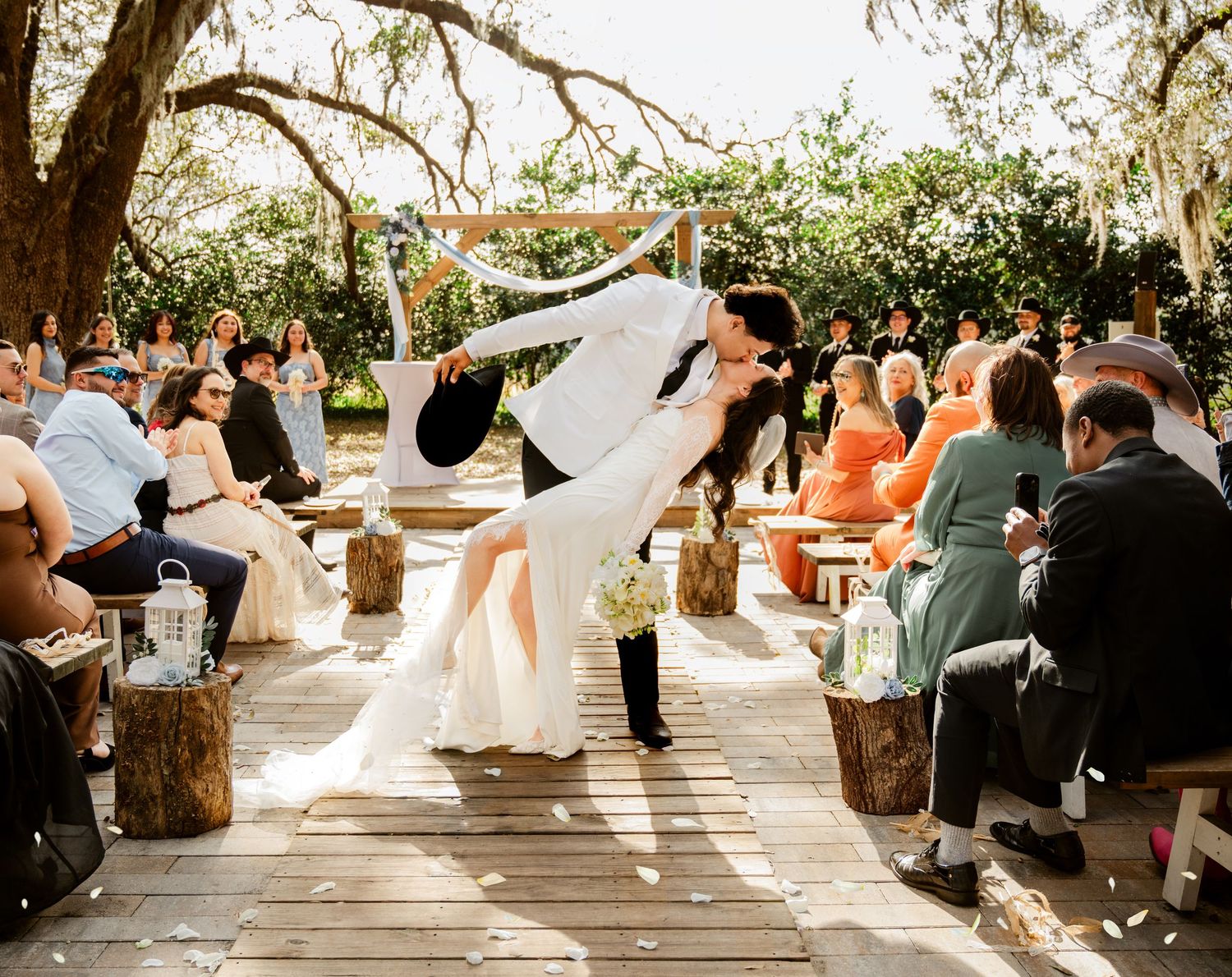 Bride and groom share a romantic dip kiss at outdoor wedding ceremony surrounded by guests on wooden benches.