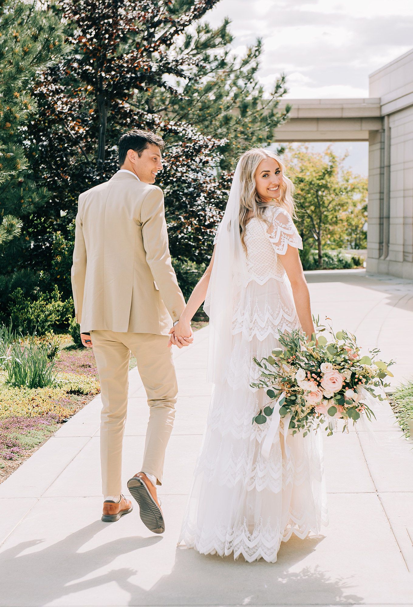 Bride in lace gown holding bouquet walks with groom in tan suit on sunny outdoor wedding day path.